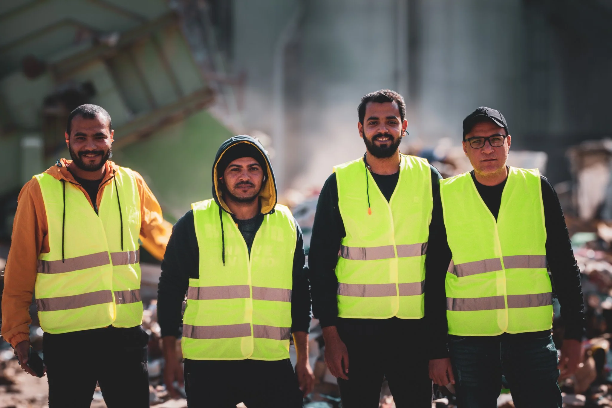 Four men wearing high-visibility safety vests standing in a landfill or waste disposal site.