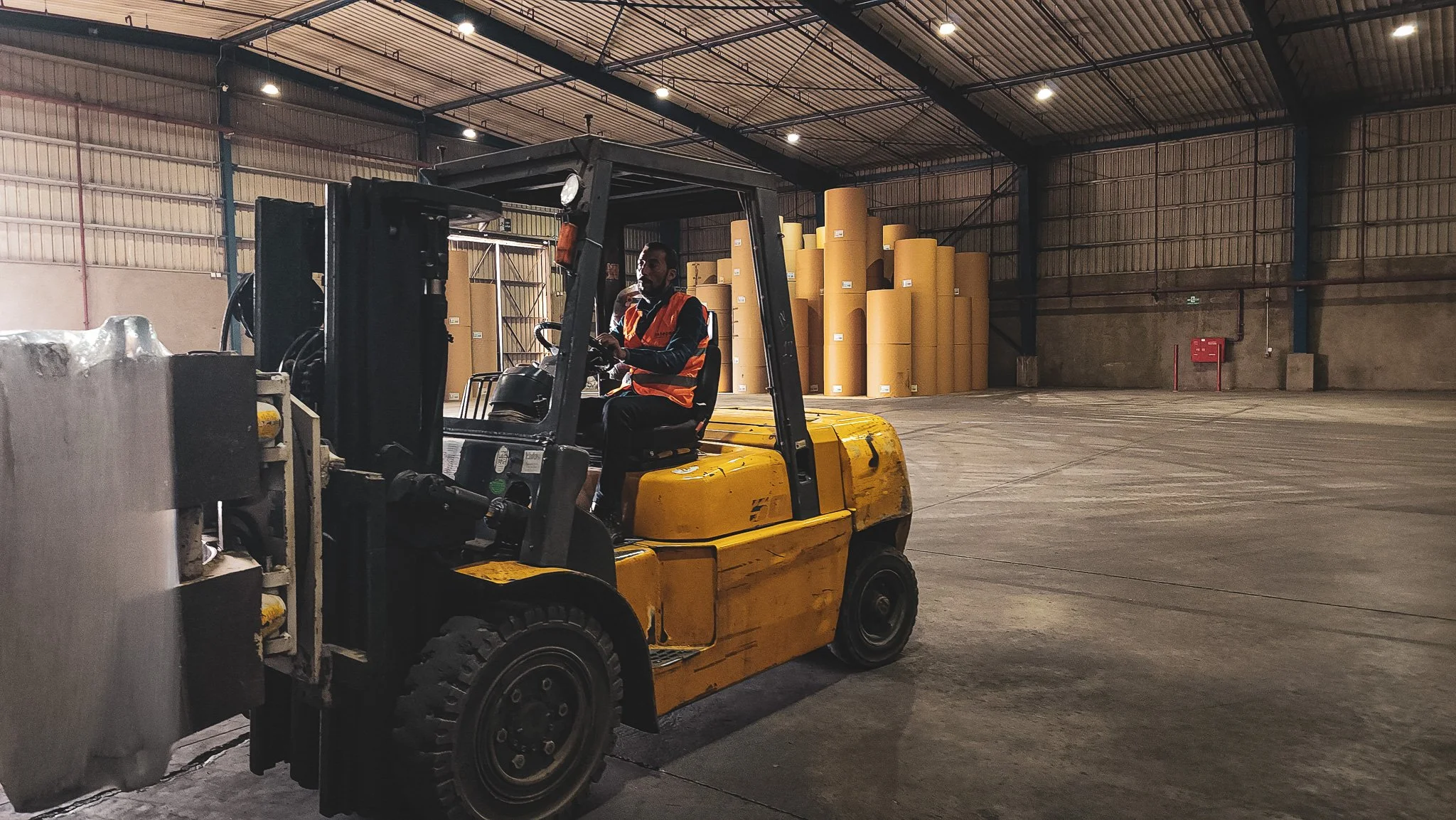 A man wearing a safety vest operates a yellow forklift inside a large warehouse with a concrete floor and high ceiling. Stacks of large yellow paper rolls are seen in the background.