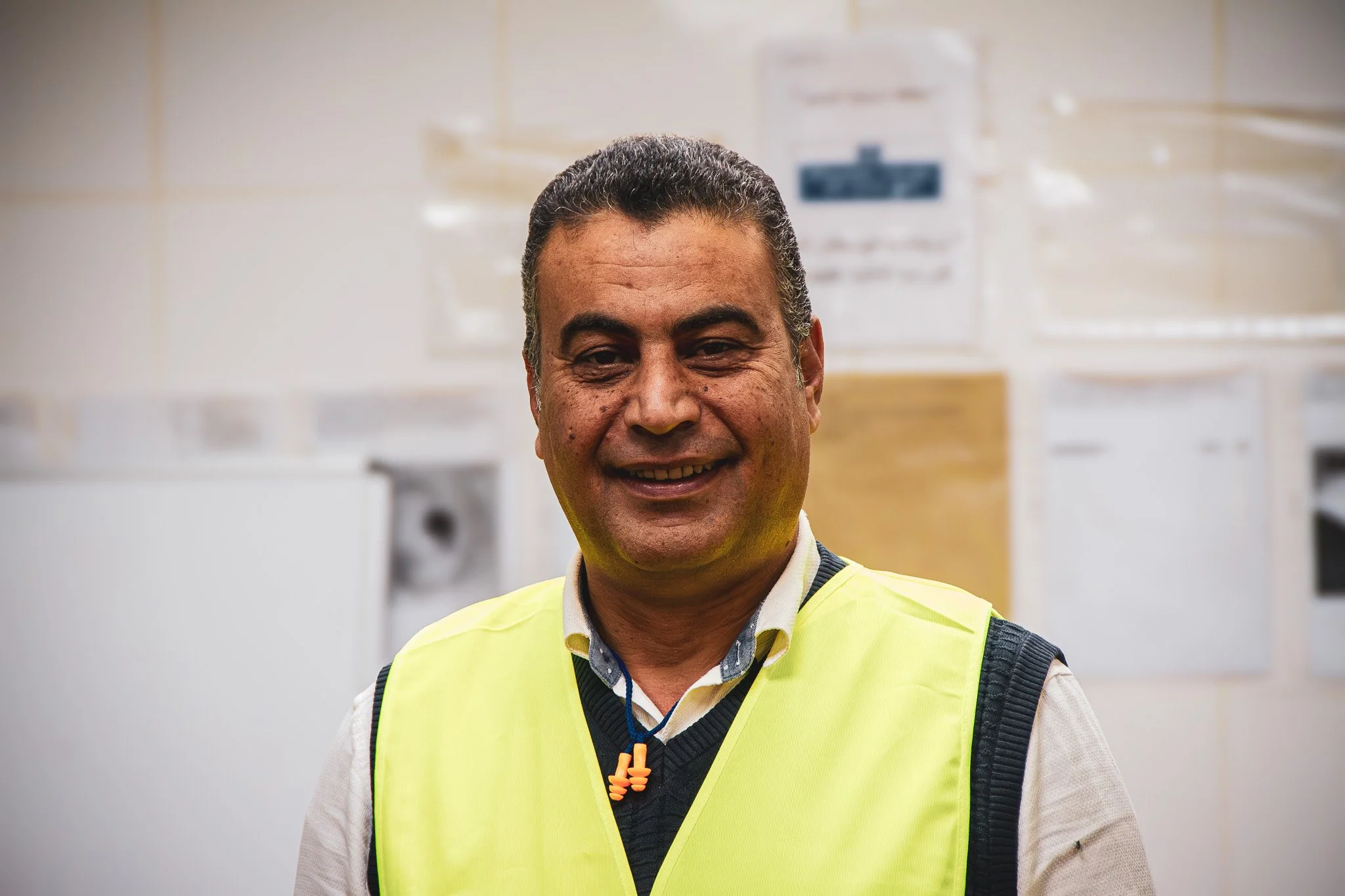 A man smiling, wearing a yellow safety vest over a dress shirt, standing in front of an off-white tiled wall with electrical panels and equipment.