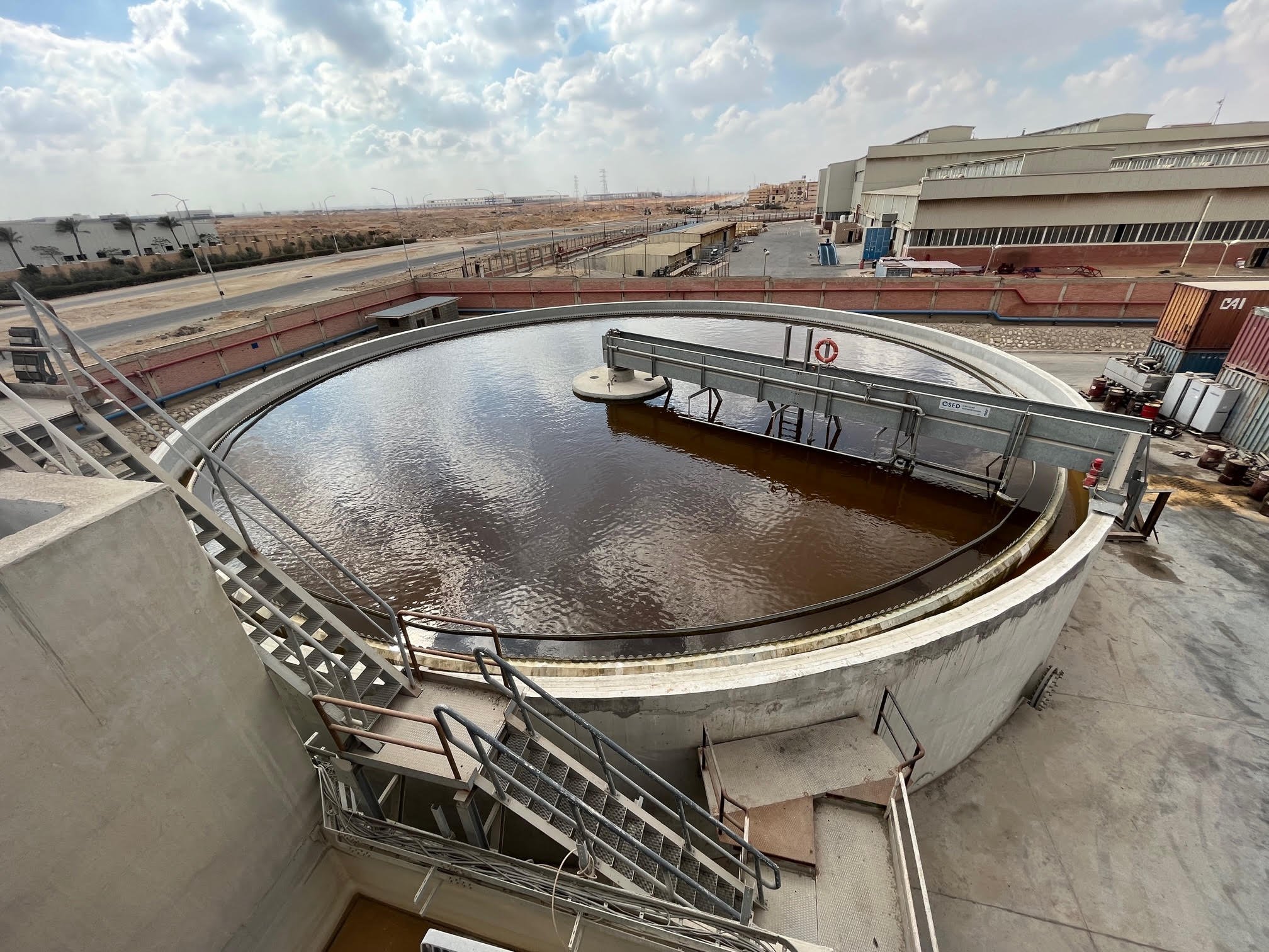 Large circular water treatment tank outdoors, with brown water and equipment around it, under a cloudy sky.