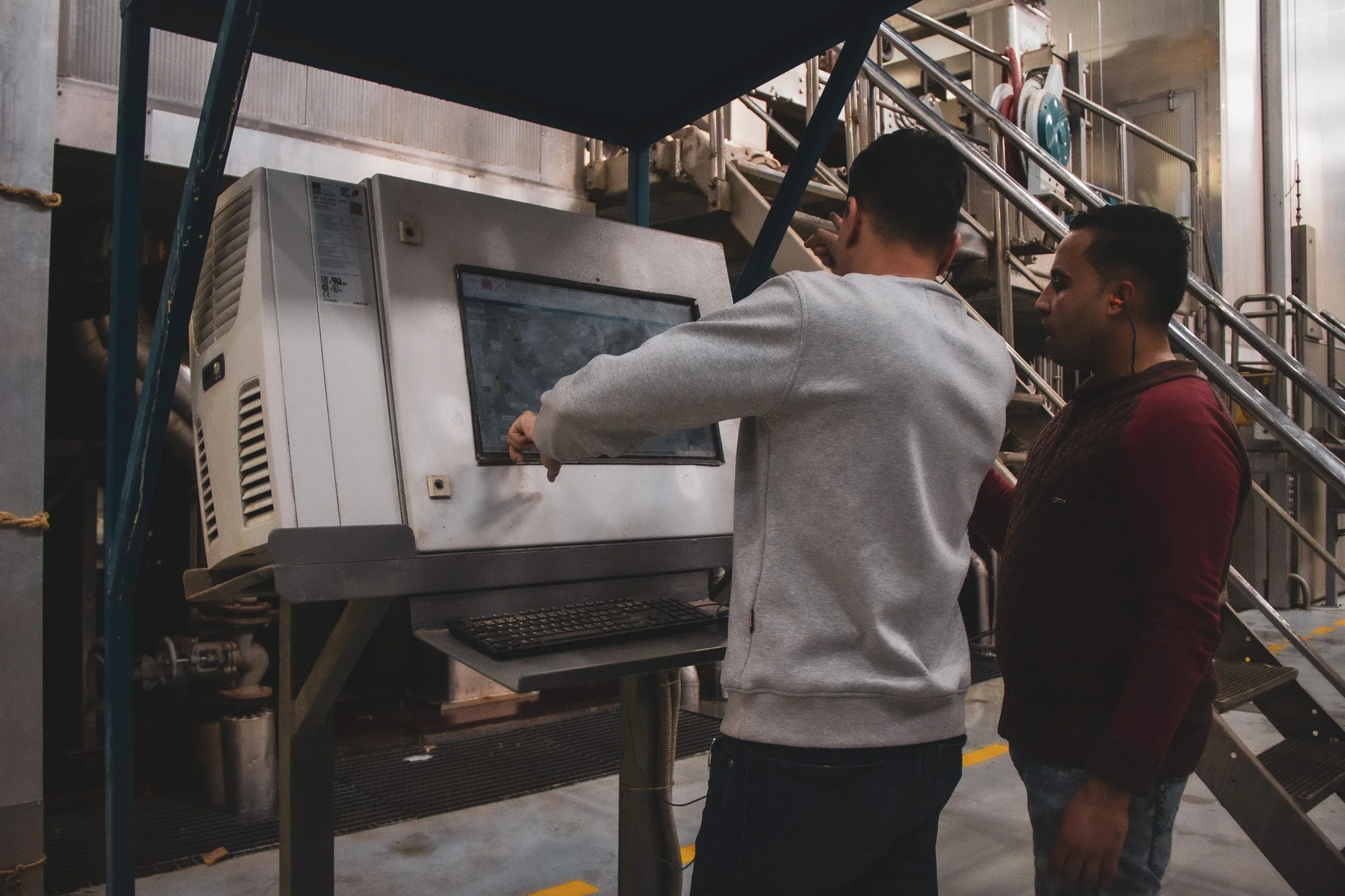 Two men work together using a touchscreen control panel in an industrial setting with metal stairs and equipment in the background.