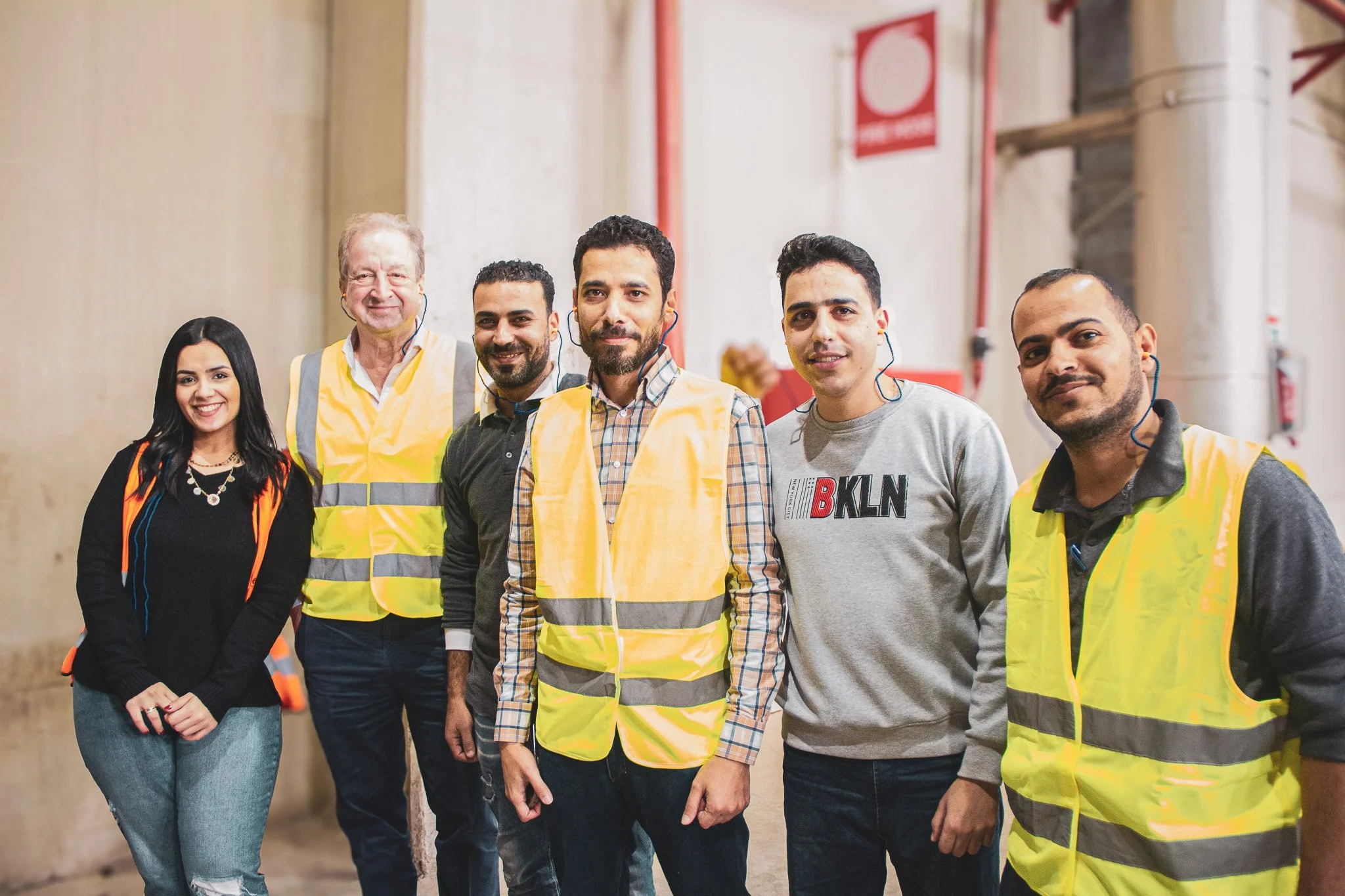 Group of six diverse workers standing in an industrial setting, some wearing safety vests, smiling at the camera.