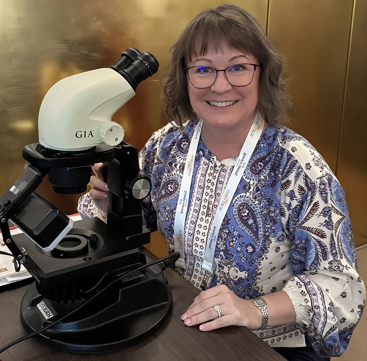 A woman with glasses and a blue patterned blouse smiling next to a microscope labeled GIA at an indoor event.