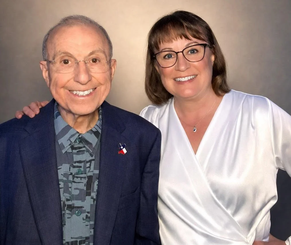 A smiling elderly man and a smiling middle-aged woman standing close together, with the woman's hand on the man's shoulder, against a neutral background.