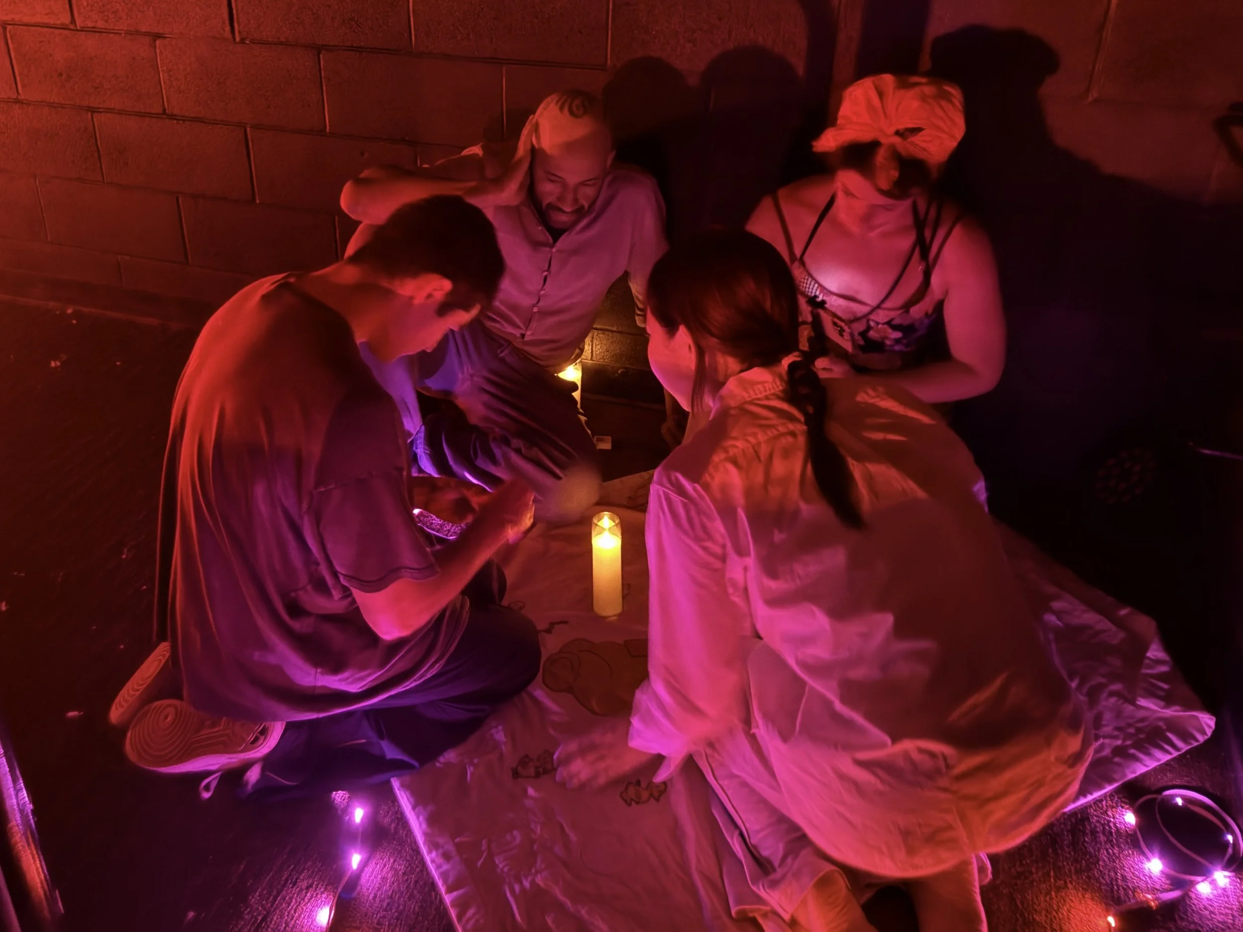 Five people sitting on the floor in a circle, engaged in a spiritual or religious ritual, illuminated by a candle and purple string lights.