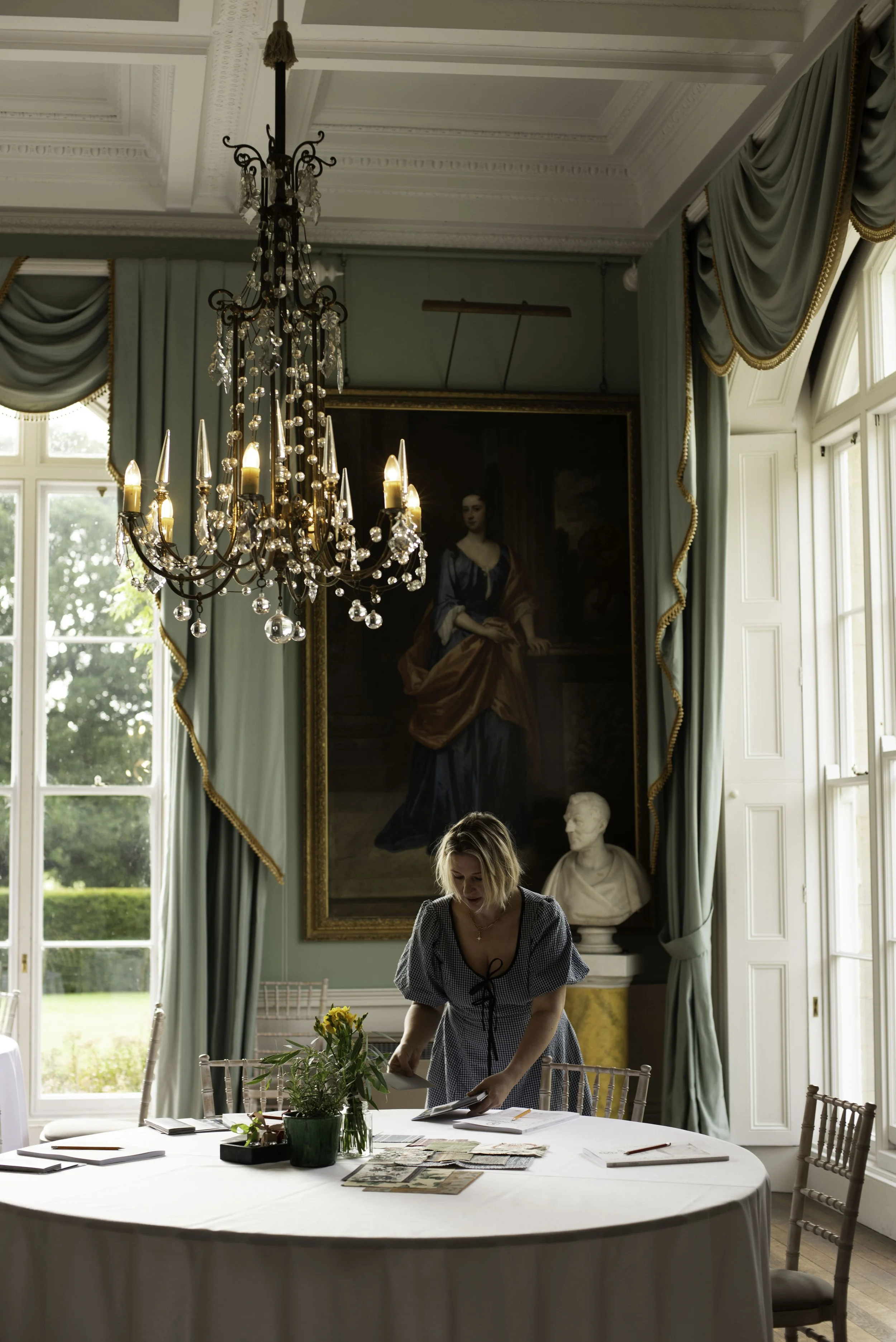 A woman in a checkered dress stands at a round table with papers, magazines, and a potted plant with yellow flowers in a large, ornate room with high ceilings, large windows, a chandelier, a portrait painting, and a bust sculpture.