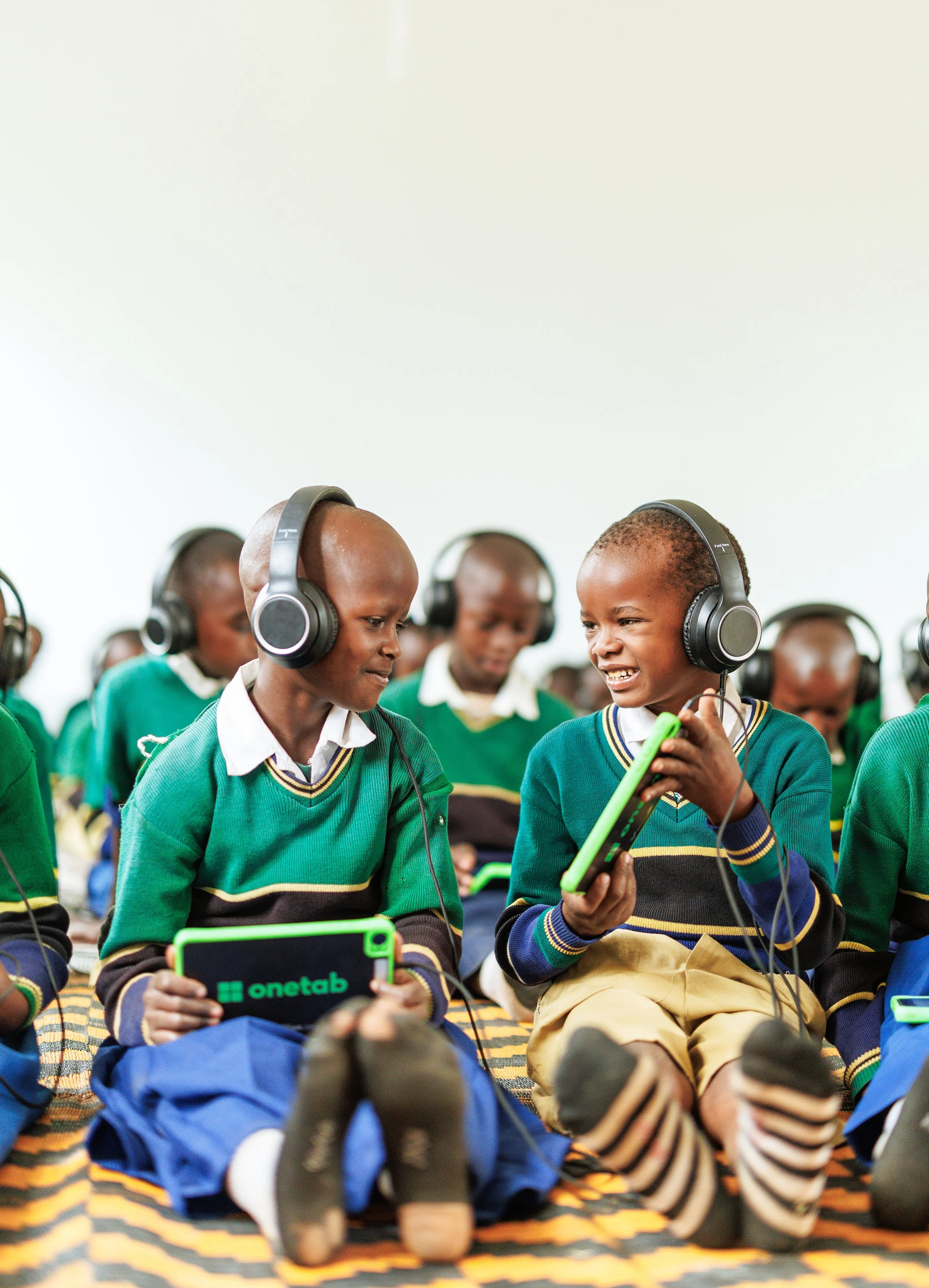 Group of school children in green uniforms with headphones, sitting on a blanket, engaging with tablets.