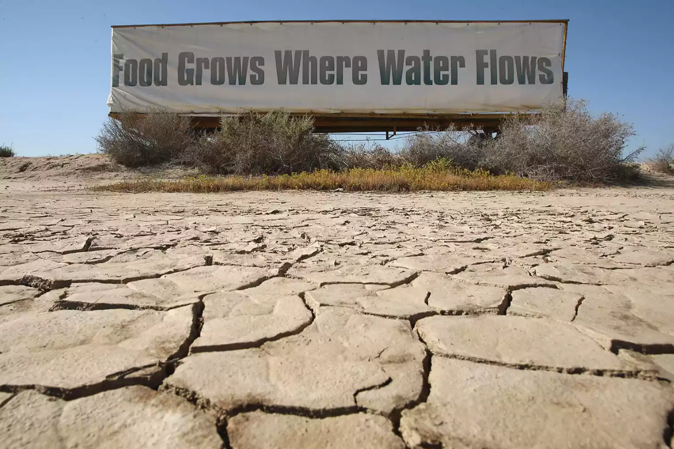 Dry, cracked desert ground with bushes and a billboard in the background reading "Food Grows Where Water Flows".