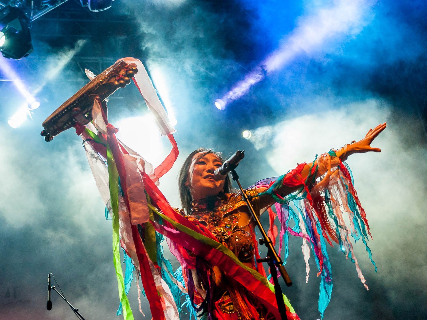 Performer on stage wearing a colorful costume with ribbons, holding a tambourine in one hand and raising the other arm, surrounded by stage lights and smoke.