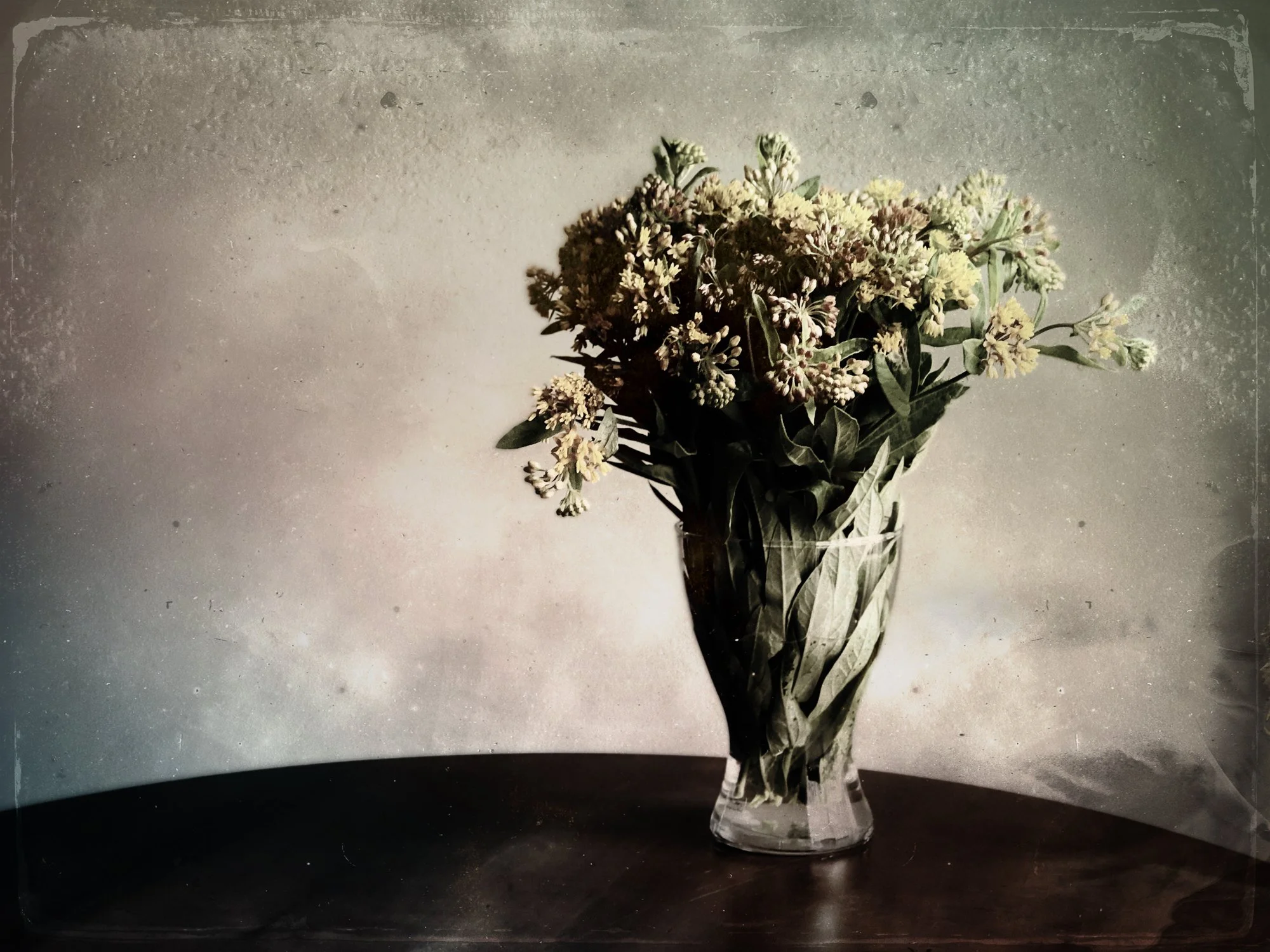 A clear glass vase with a bouquet of white and light yellow flowers and green leaves placed on a dark surface with a plain textured background.
