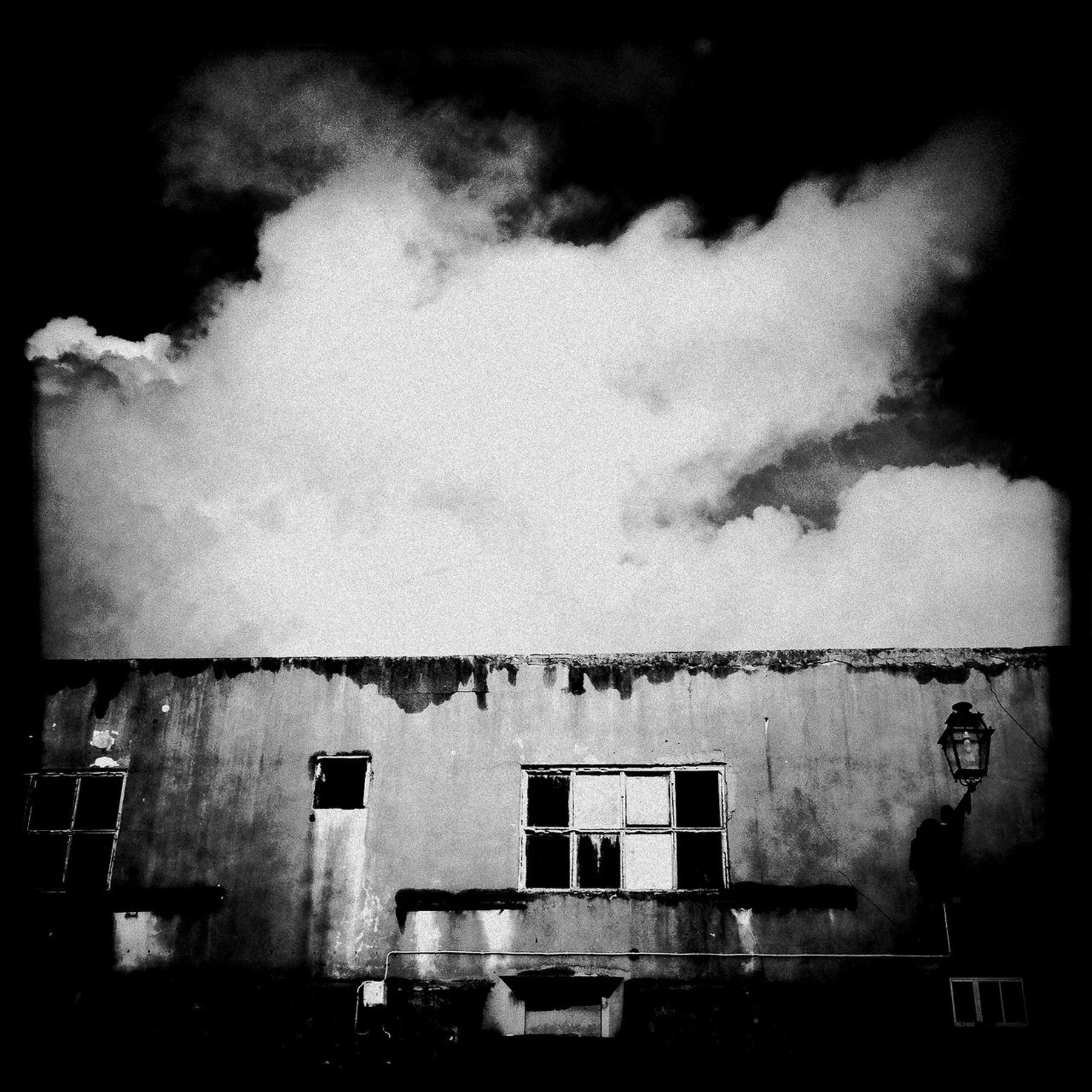 Black and white photo of a weathered building with broken windows and a street lamp, with dark clouds overhead.