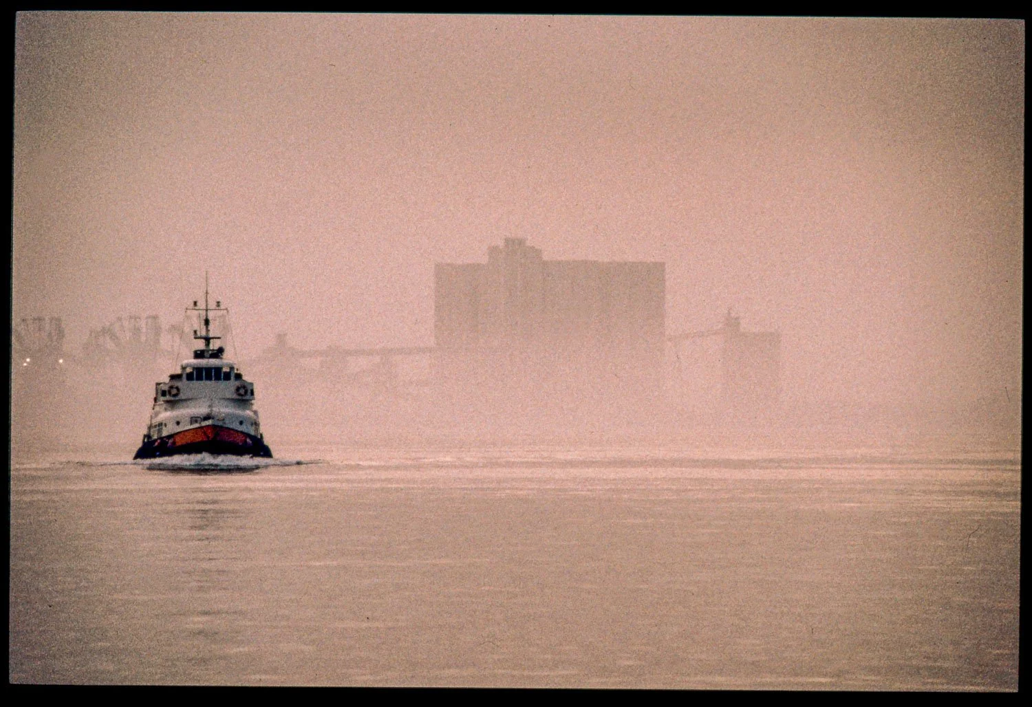 A boat sailing on calm water with a city skyline and high-rise buildings in the background, shrouded in fog or haze.