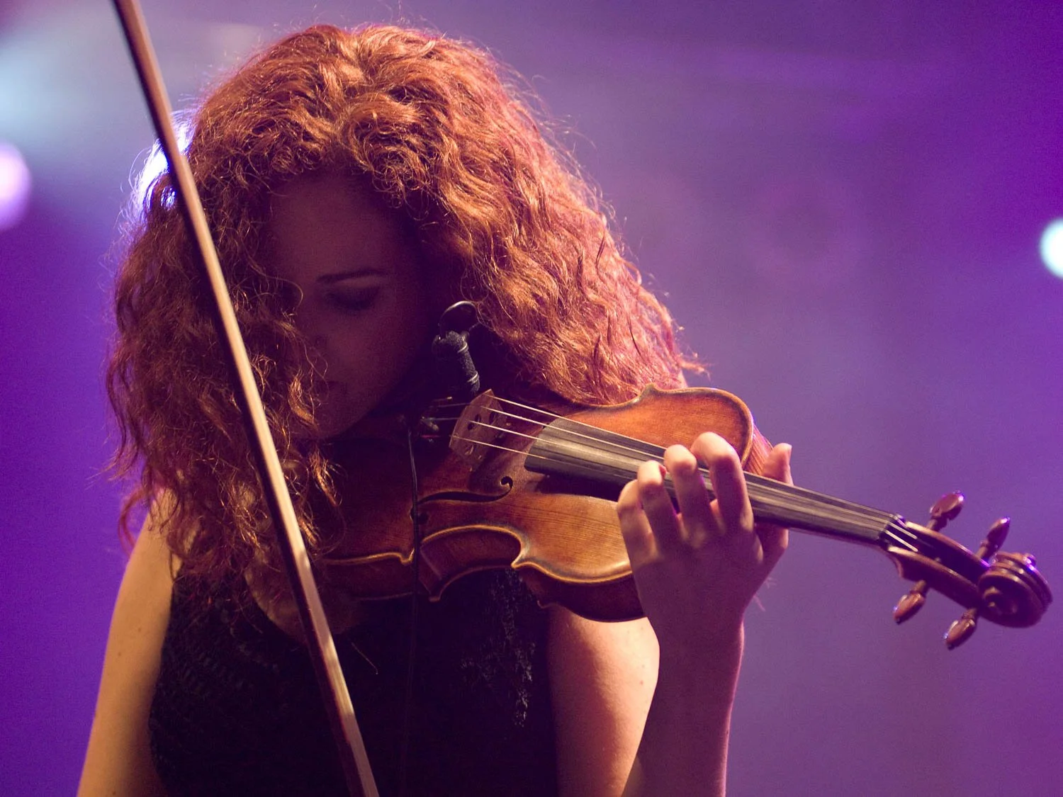 A woman with curly red hair playing the violin on stage, with purple and blue stage lighting