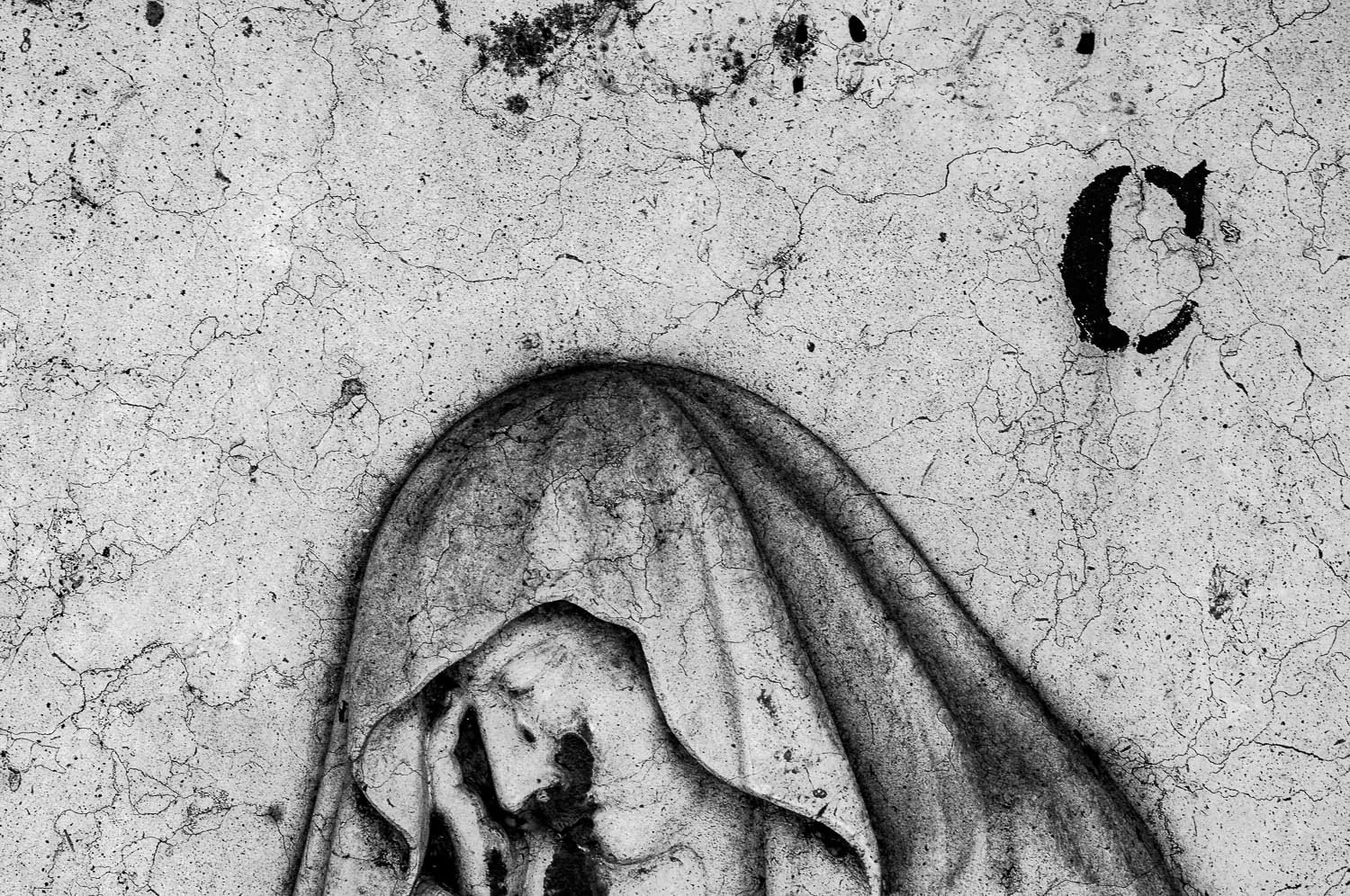 A black and white photograph of a weathered wall with a religious mural of a veiled woman holding a crying face, possibly depicting Mary with the Christ child, and the letter 'C' carved or painted on the wall.
