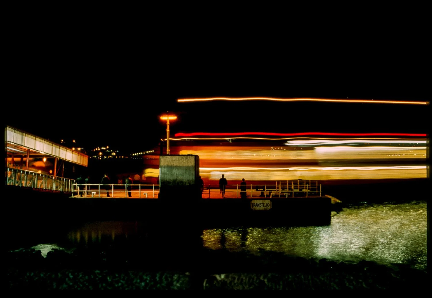 Nighttime scene of a pier with people standing on it, and light trails from boats or ships passing by reflected in the water.