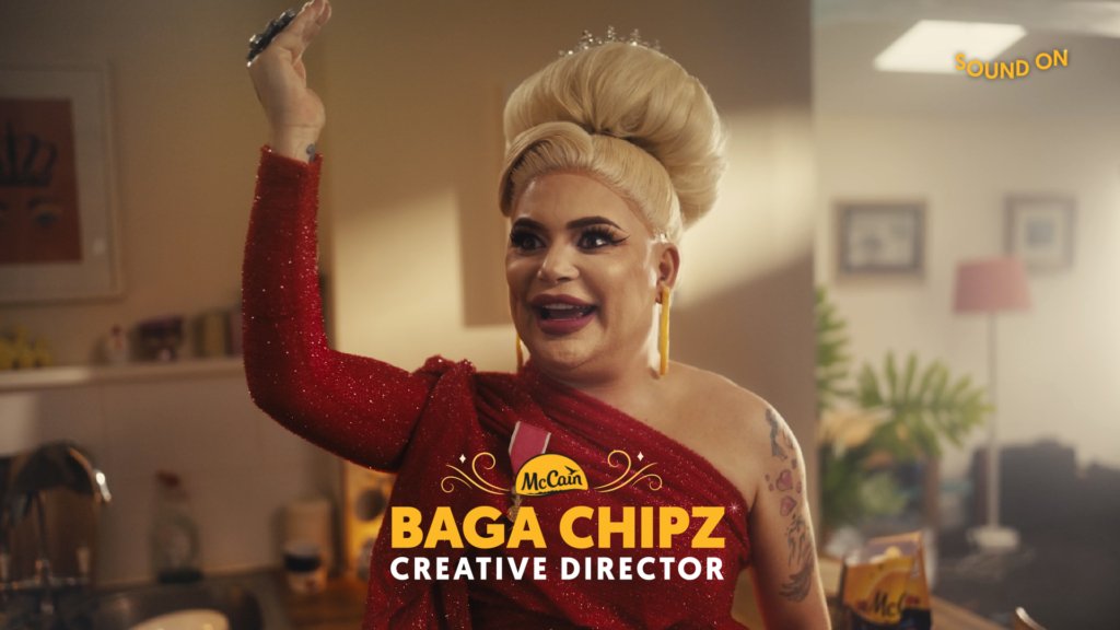 Drag Queen Saga Chipz stands in an apartment and looks into the distance with one hand raised.  