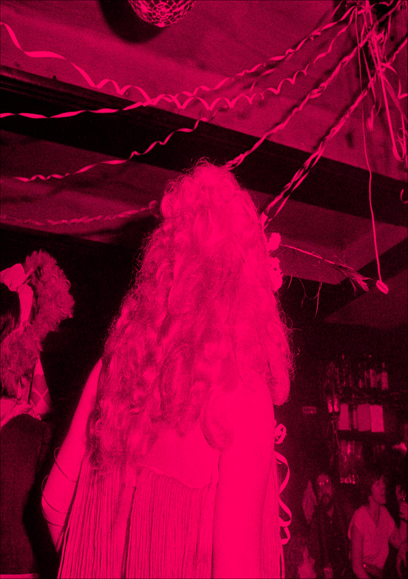 A pink-tinted image of a crowded indoor party scene with streamers and people dancing, one person with long curly hair in the foreground.