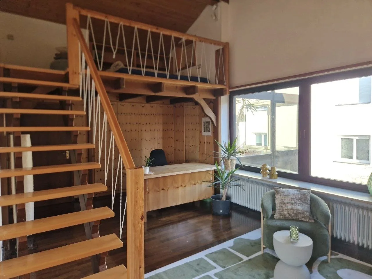 Gemütliches Wohnzimmer mit Holztreppe, Fenster, grüne Sessel, Pflanzen, Couchtisch, Teppich und Schreibtisch.