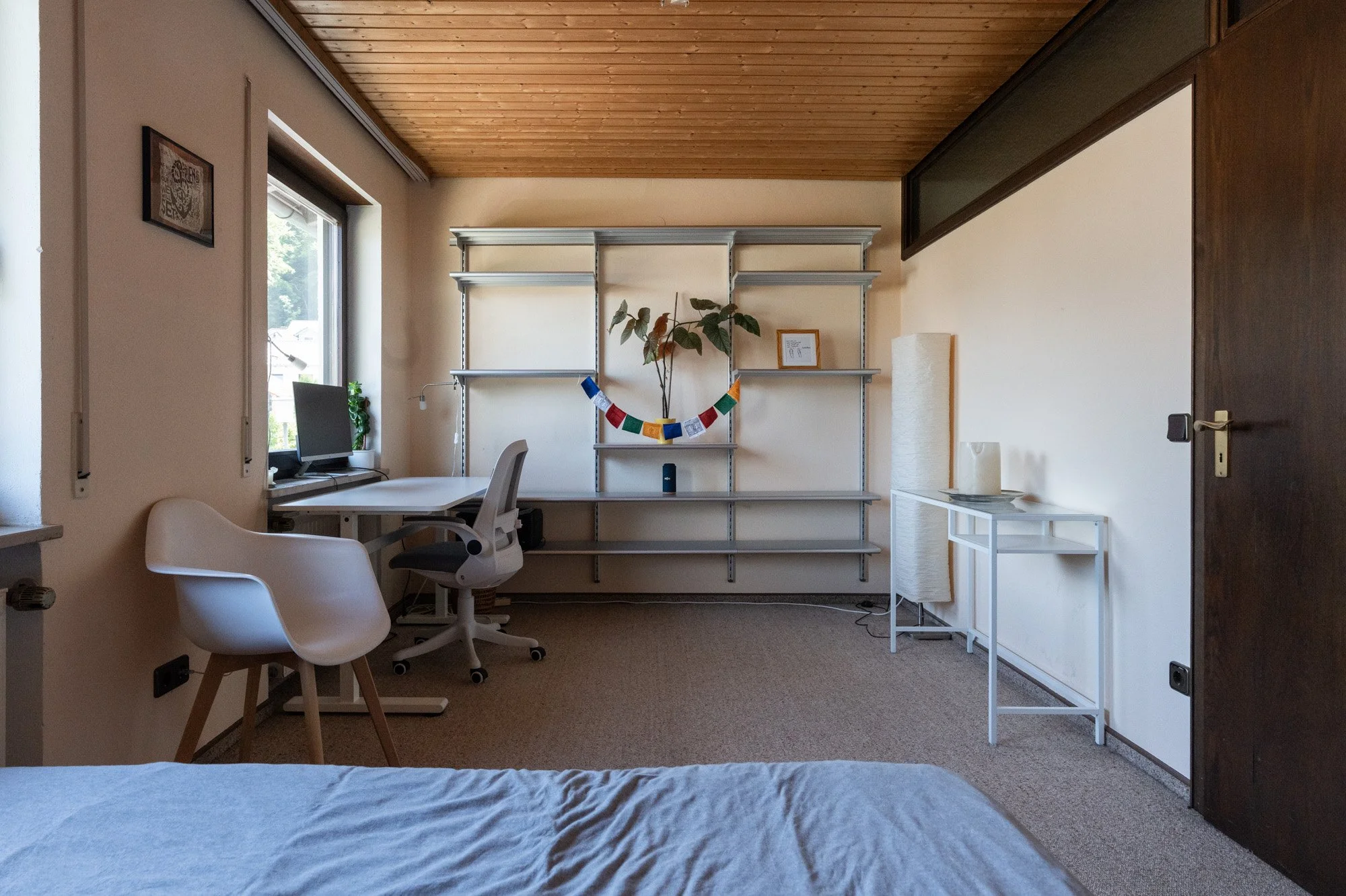 Schlafzimmer mit Bett, Schreibtisch, Monitor, Bürostuhl, Bücherregal, Pflanze, Stehlampe, Zimmerdeko