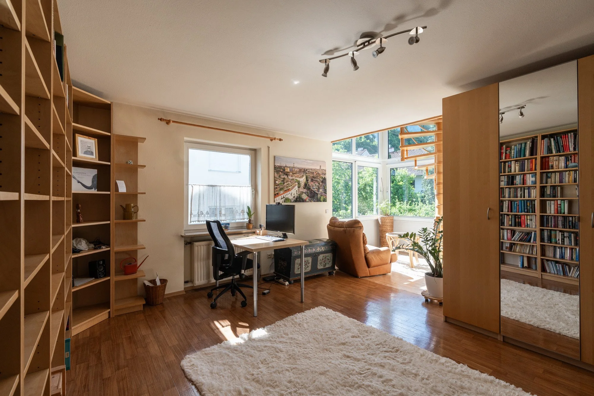 Wohnzimmer mit Holzfußboden, weißen Wänden, großen Fenstern, Bücherregalen, einem Schreibtisch mit Stuhl, einem Sessel, einem Fernseher, Pflanzen, einer weißen Teppich und einem Wandbild.