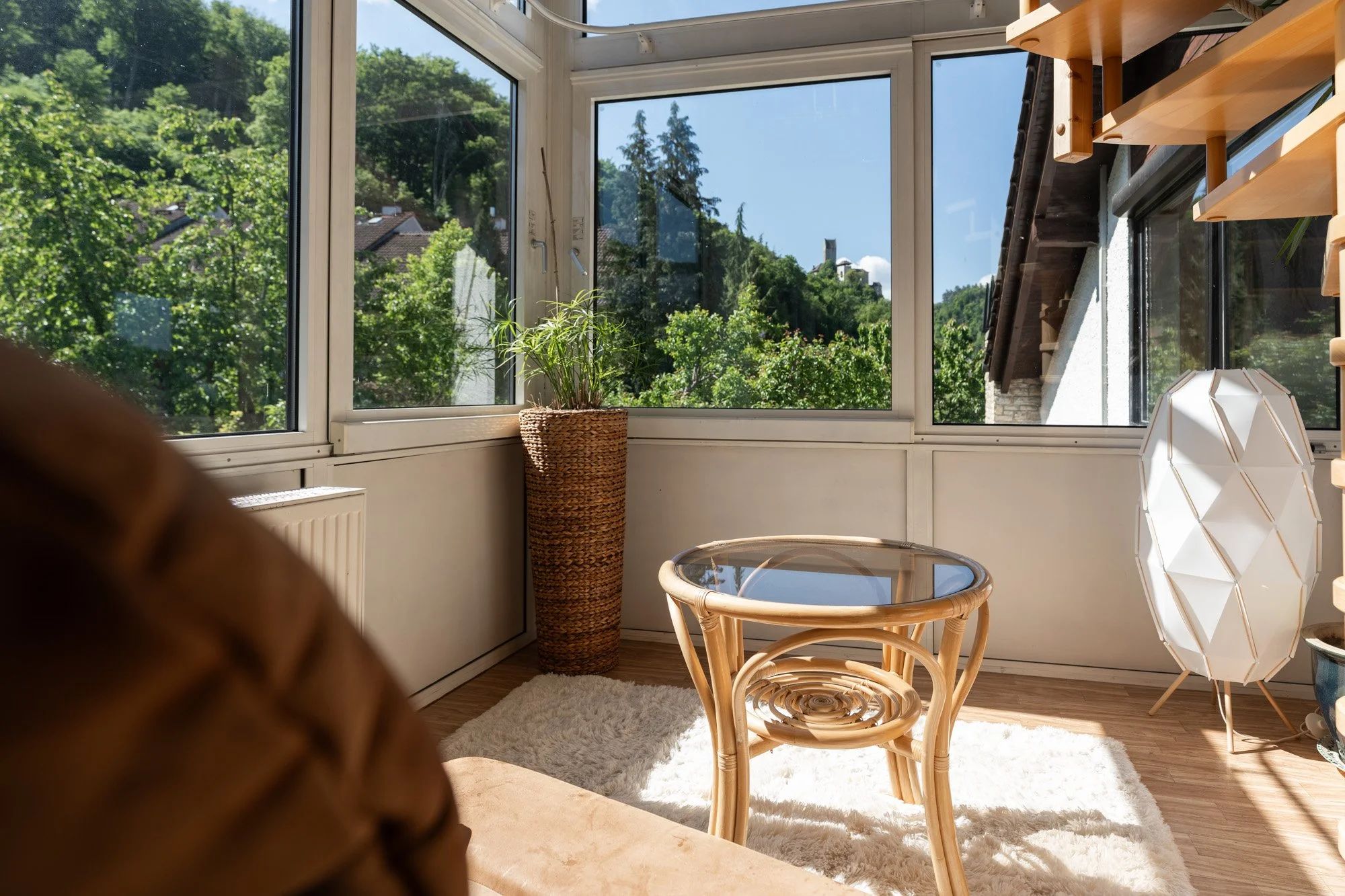 Helles, sonniges Wohnzimmer mit großen Fenstern, einer Pflanze in einem hohen Korb, einem Rattanstuhl mit Glastisch, Teppich und Stehlampe.