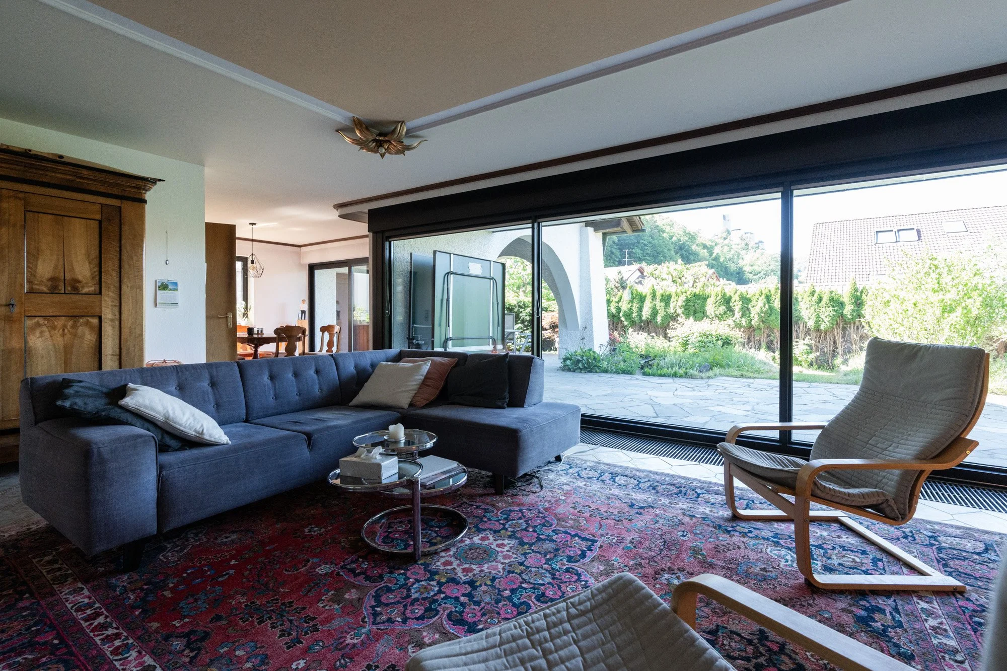 Gemütliches Wohnzimmer mit dunkelblauer Couch, Holzarmlehnenstuhl, buntem Teppich, großen Fenstern mit Blick ins Freie.