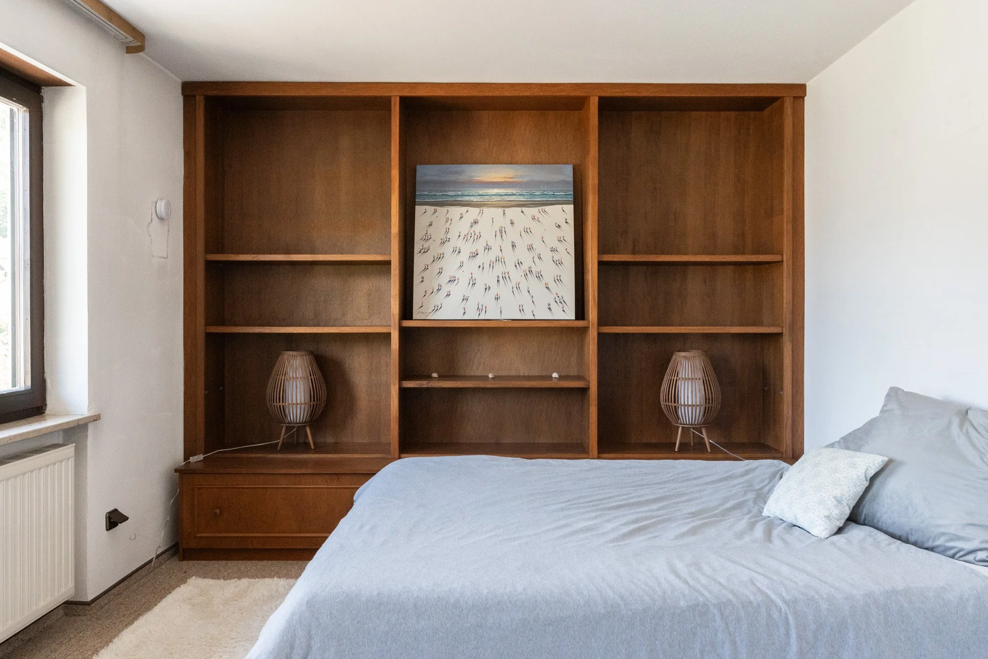 Schlafzimmer mit Holzwandregal, Bett mit hellblauer Bettwäsche, zwei Lampen und einem Wandbild eines Strandes mit Menschen, an einem Fenster mit weißen Wänden und einem Heizkörper.