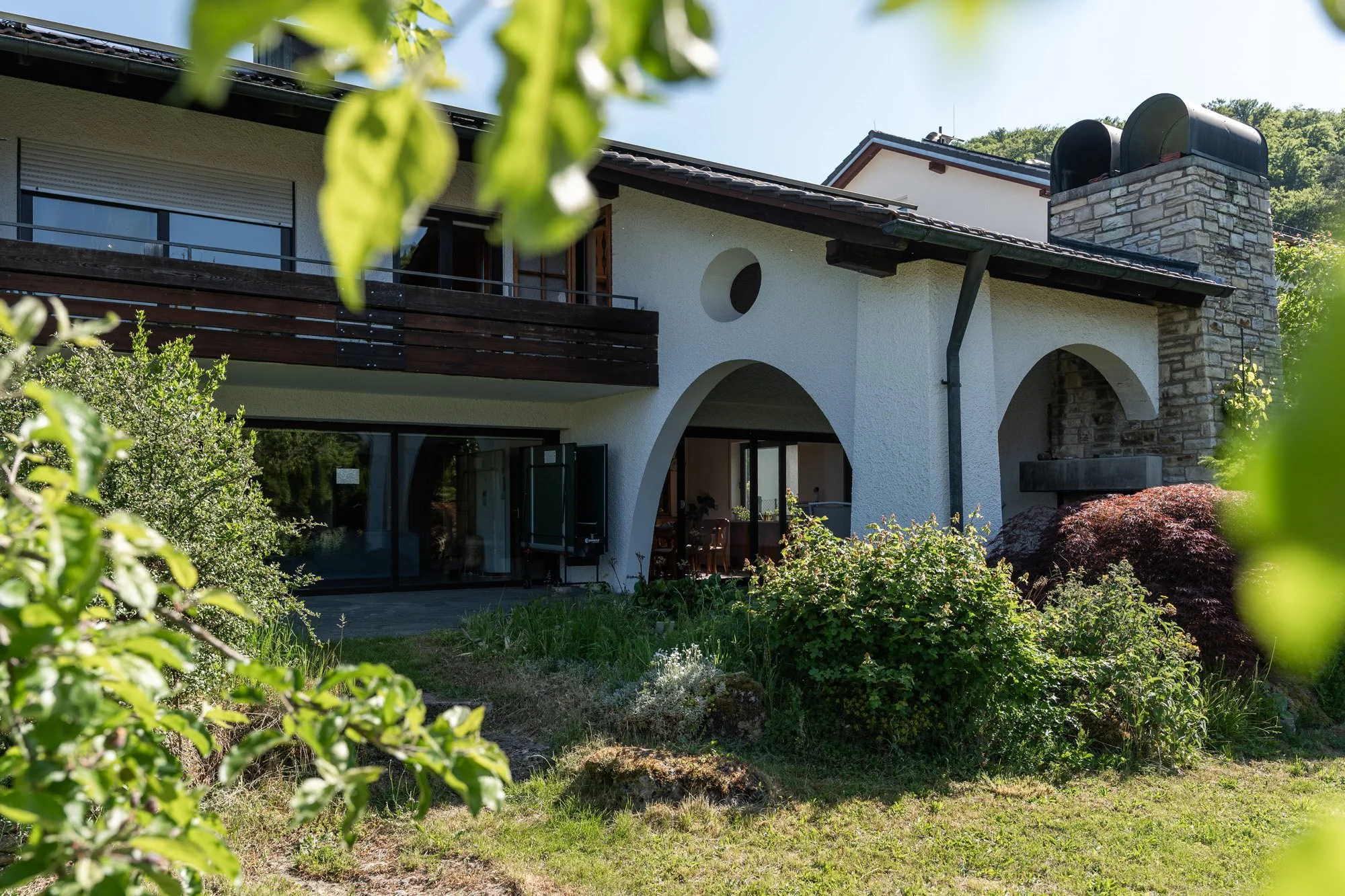 Ein Haus mit weißer Fassade, holzverkleckter Balkon, große Fenster und Gehwege, umgeben von Büschen und Bäumen an einem sonnigen Tag.