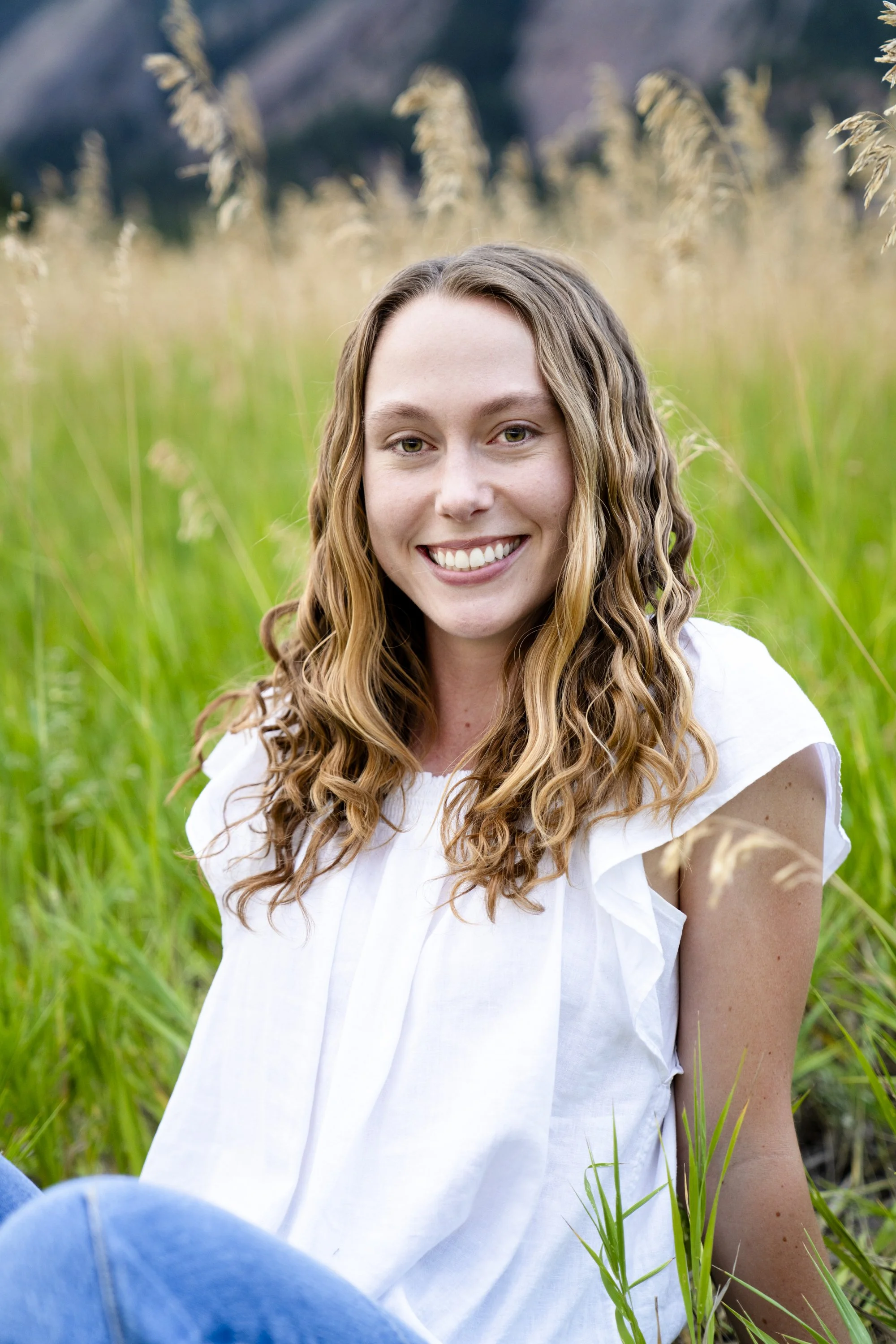 A young woman with wavy blonde hair smiling while sitting in a grassy field with mountains in the background.