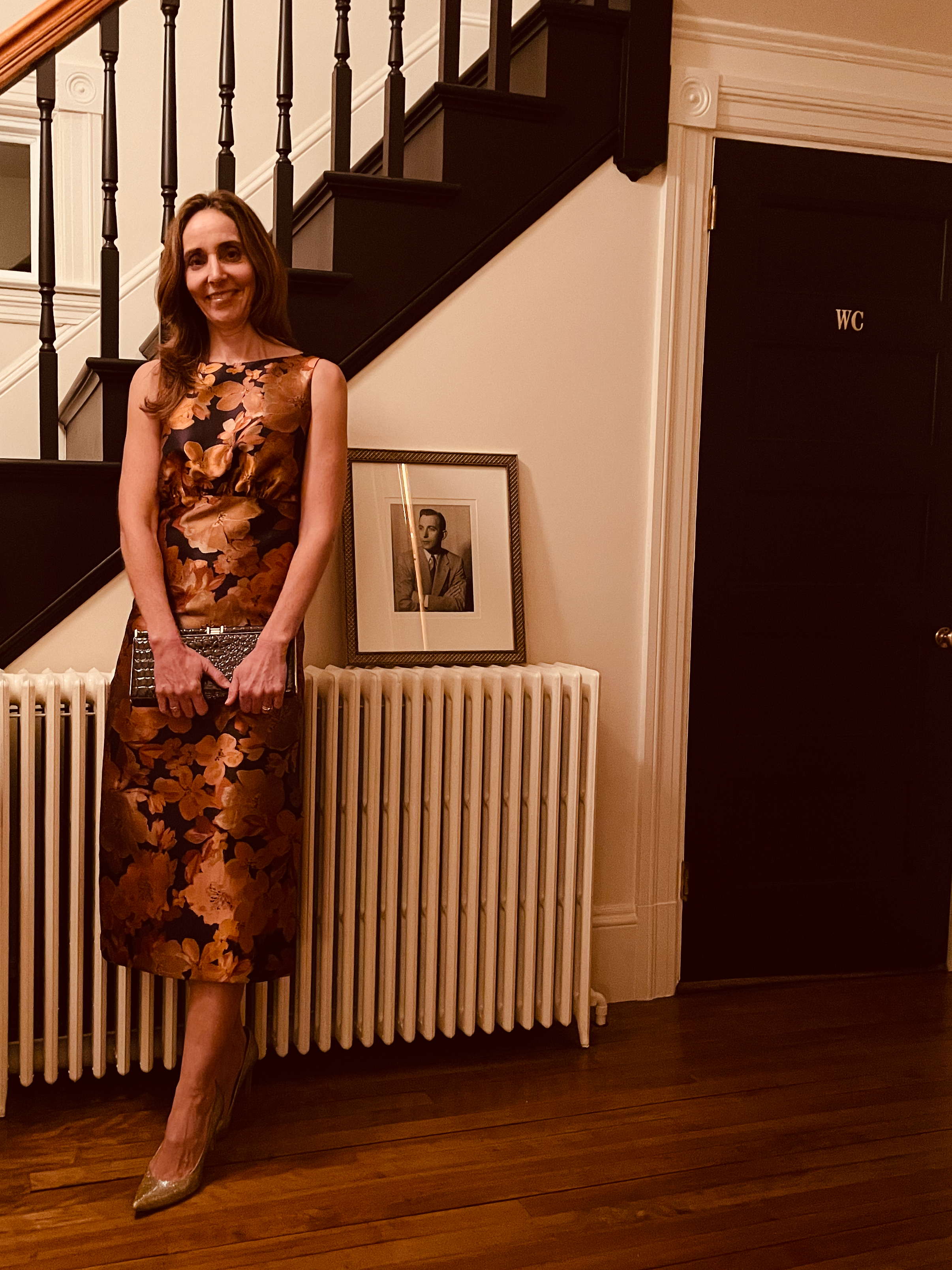 A woman in a floral dress standing indoors against a radiator, holding a clutch, with a framed black-and-white photograph on the radiator and a staircase behind her.