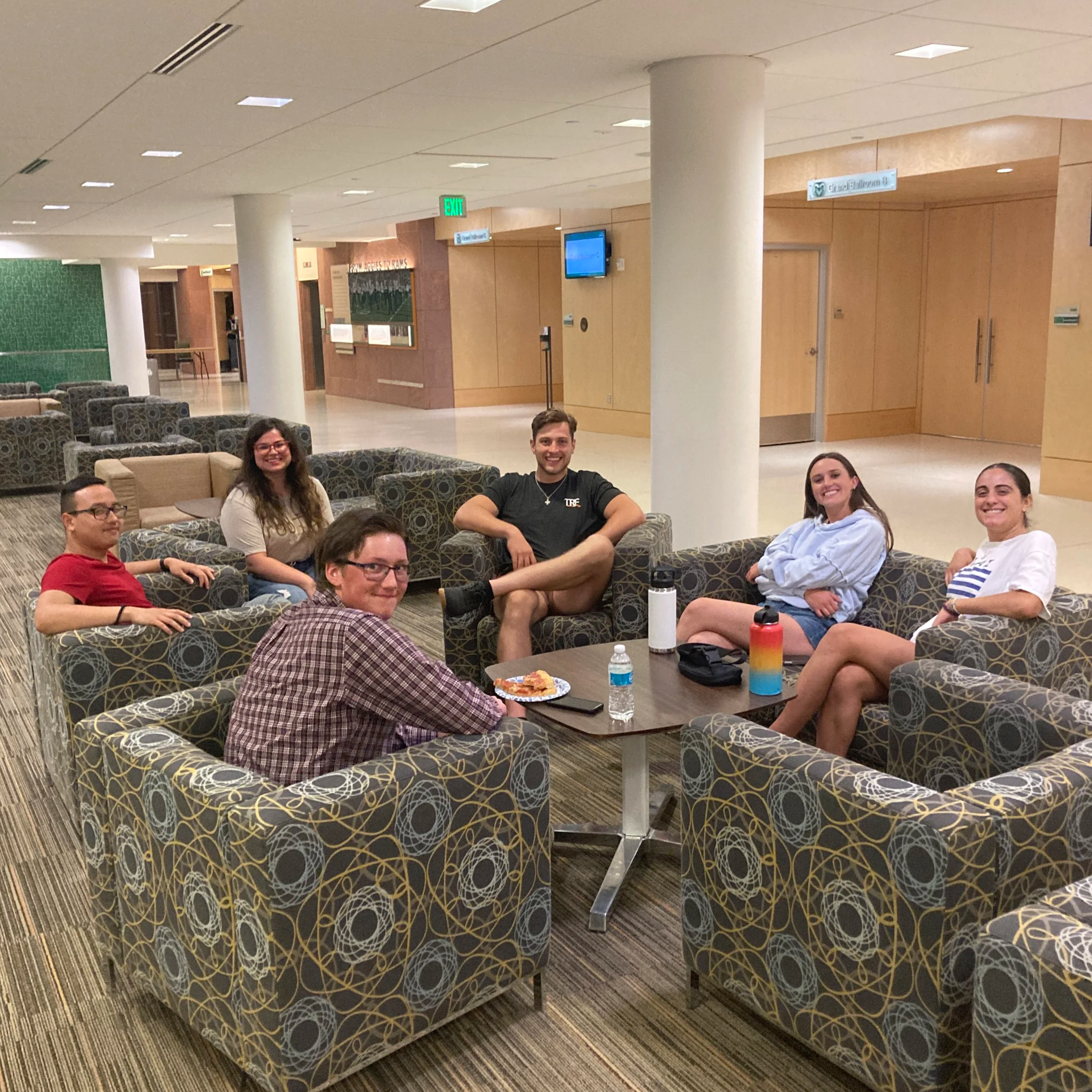 Group of six people sitting in a lounge area with patterned armchairs and a table. They appear relaxed and are smiling. The background shows a hallway with signs, doors, and a display screen.