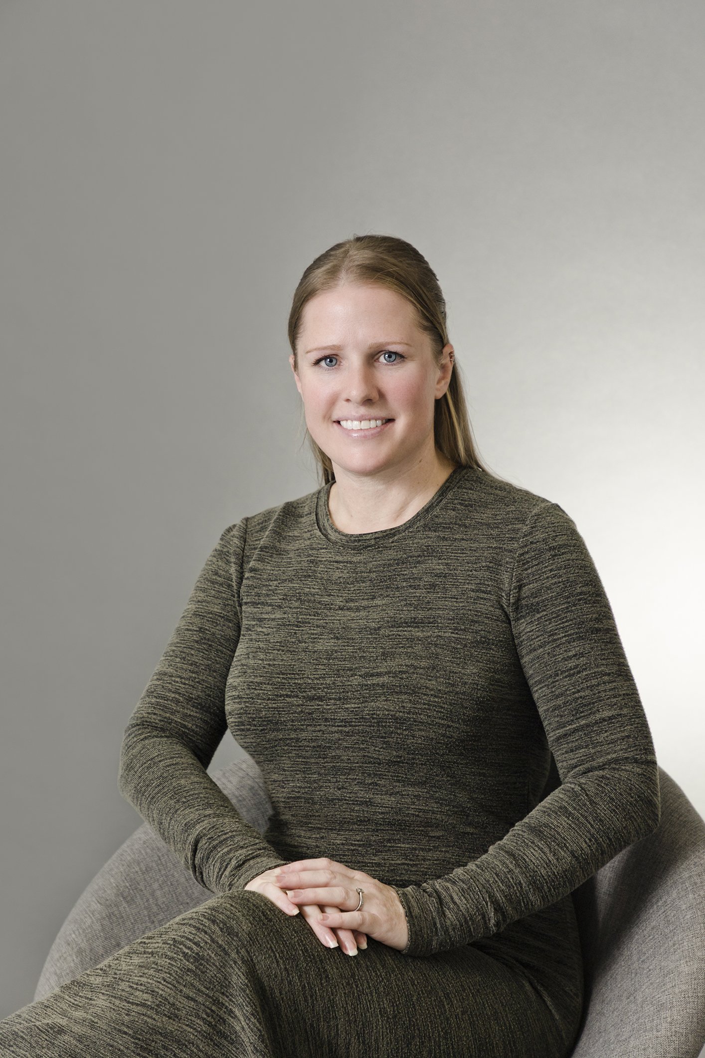 A woman with light brown hair, wearing a dark green, long-sleeved dress, sitting on a gray chair with a plain light gray background.