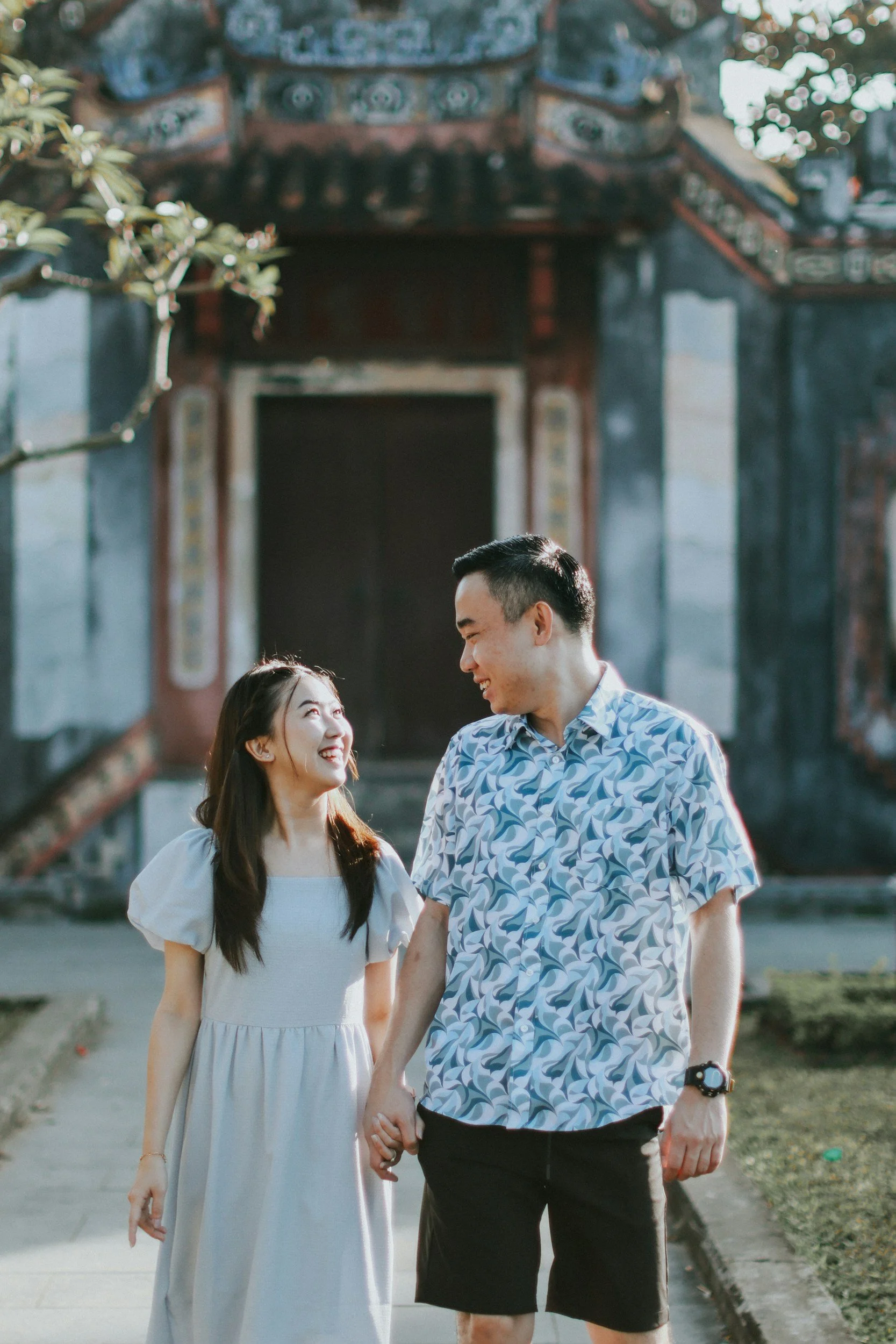 A couple holding hands and smiling at each other outdoors in front of a traditional Chinese temple.