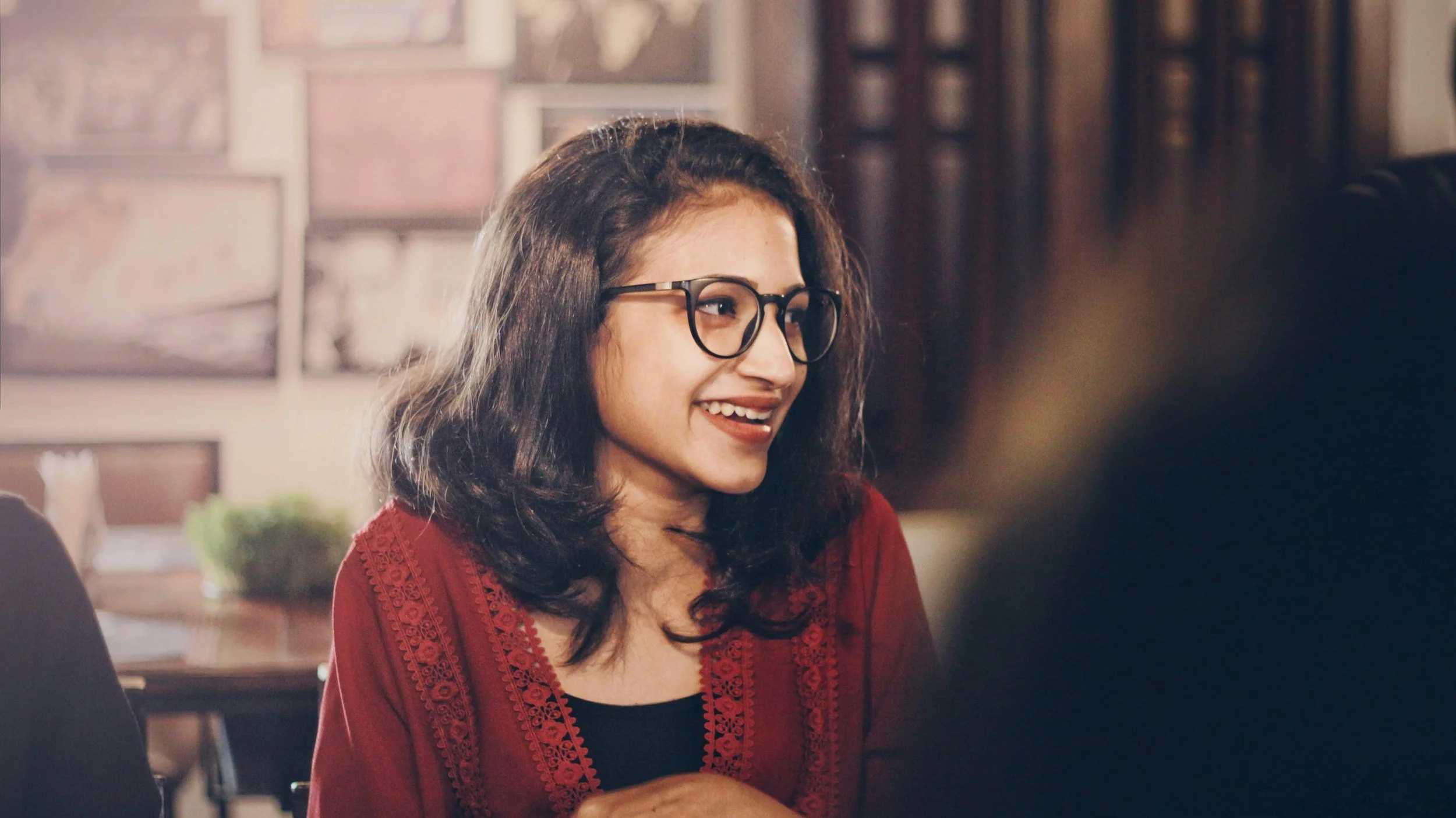 A young woman with glasses and wavy dark hair smiling in a restaurant or cafe setting.