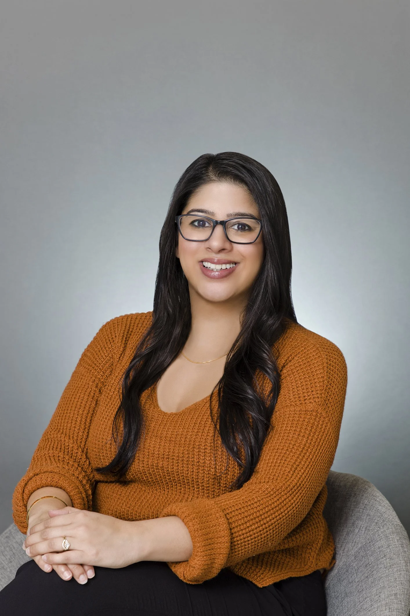 A woman with long dark hair, glasses, and a warm smile, wearing an orange sweater, sitting with her hands folded on her lap against a plain gradient background.