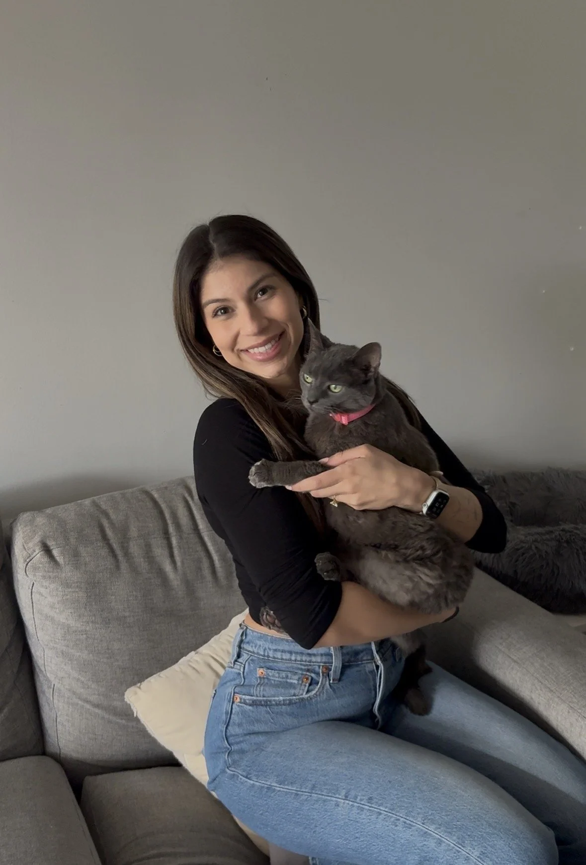 A woman with long brown hair, wearing a black top and blue jeans, is sitting on a gray couch. She is smiling and holding a gray cat with a pink collar. The woman is wearing a watch on her left wrist.