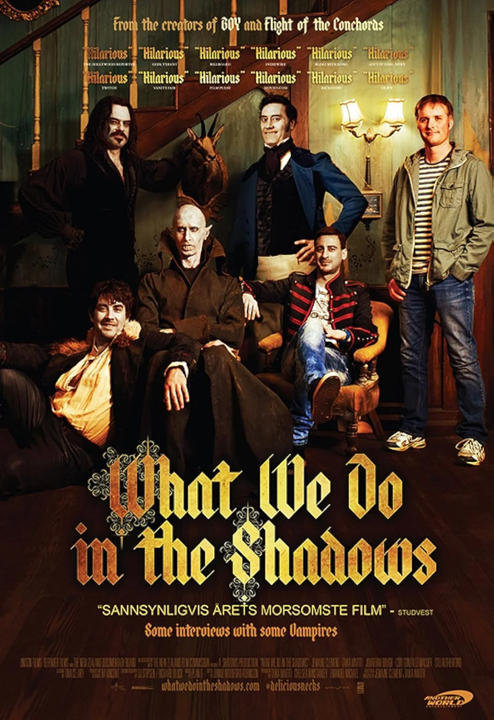 Group photo of six people and a puppet of a vampire sitting on a chair in a cozy, vintage living room. The background has a staircase, green walls, and a sign reading 'from the creators of 60s and flight of the Conchords.' The scene is from a humorous vampire-themed show or movie titled 'What We Do in the Shadows.'