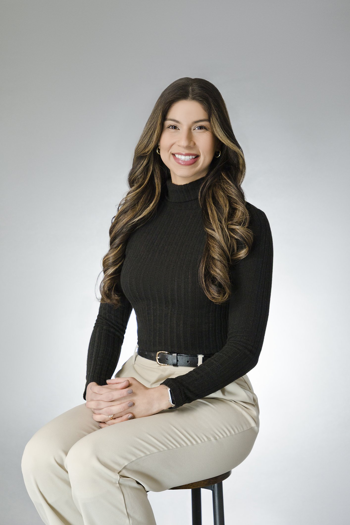 A woman with long wavy brown hair, wearing a black turtleneck and beige pants, sitting on a stool against a plain light background and smiling at the camera.