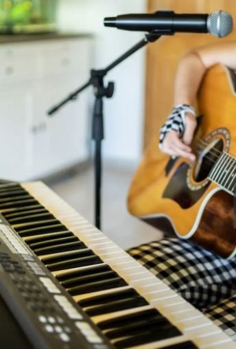 Close-up of a keyboard, a person playing an acoustic guitar, and a microphone on a stand, in a room with natural light.