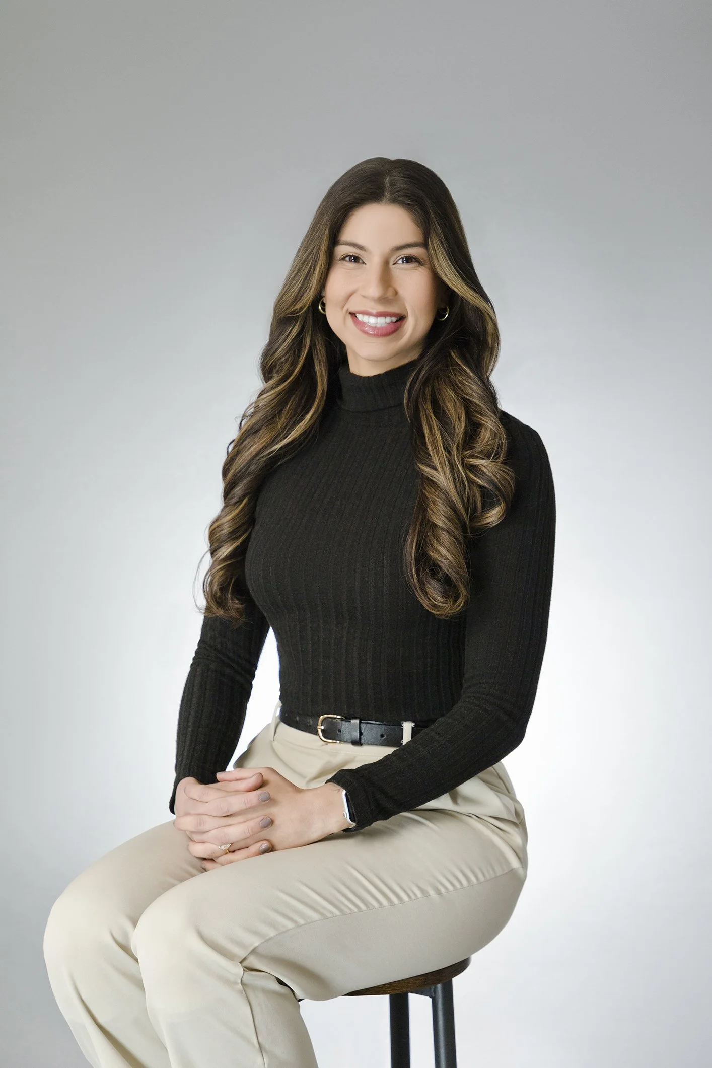 A young woman with long wavy brown hair, wearing a black turtleneck and beige pants, sitting on a stool against a plain gray background, smiling at the camera.
