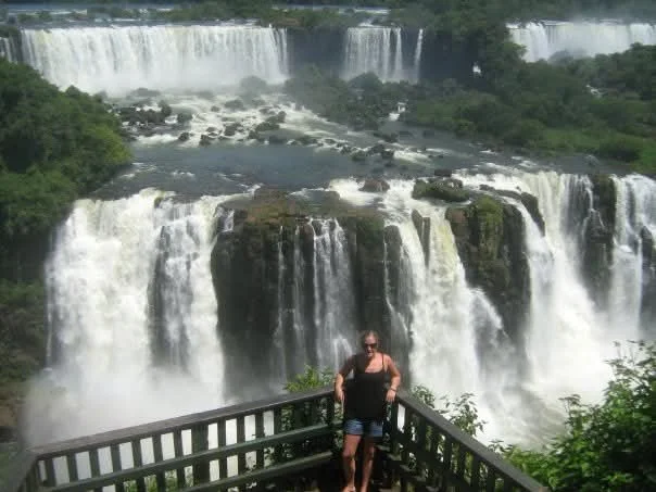 Person standing on a viewing platform overlooking Iguazu Falls with multiple waterfalls and lush green scenery in the background.