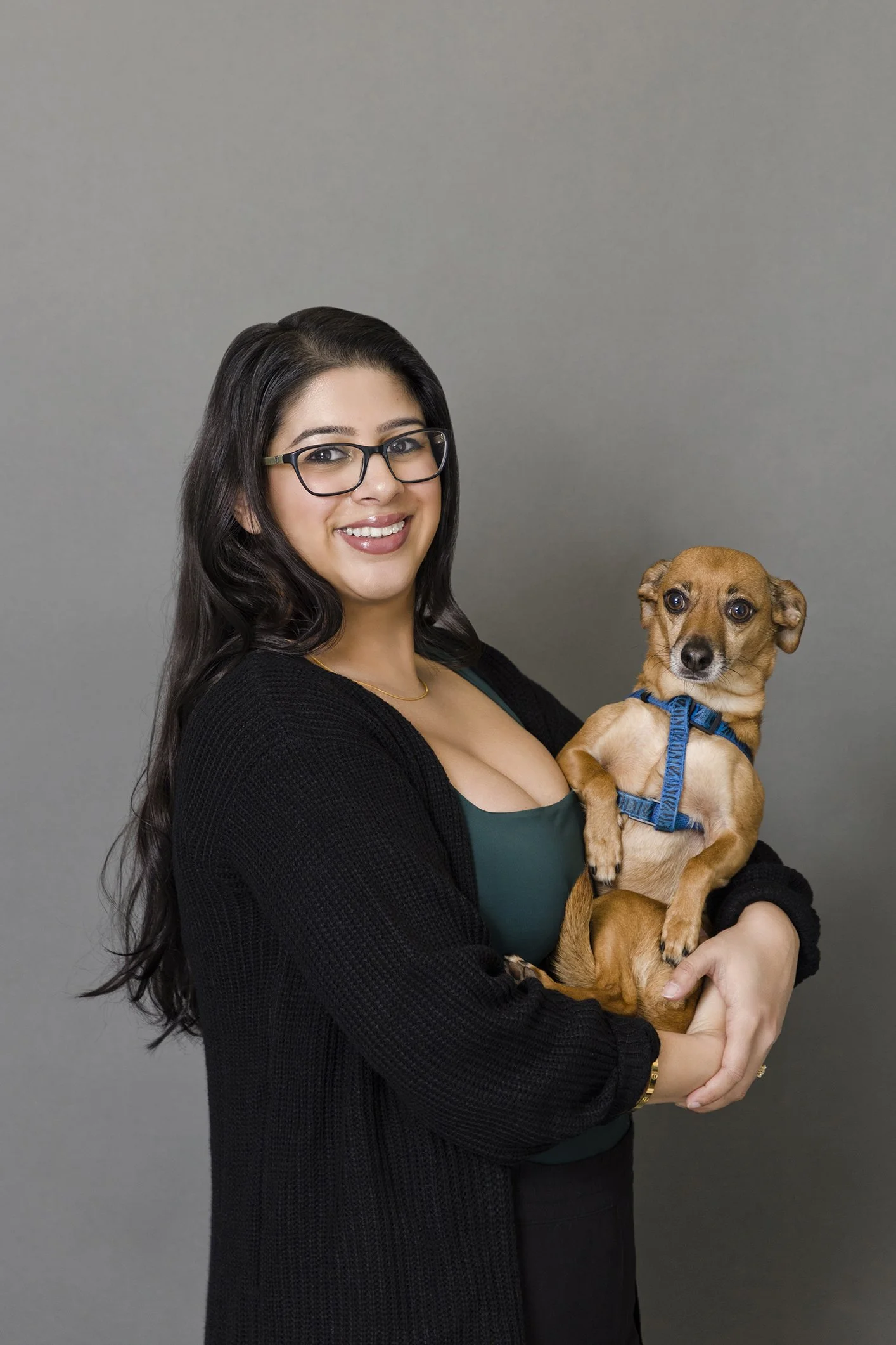 A woman with long dark hair, glasses, and a black cardigan holding a small brown dog with a blue harness against a gray background.