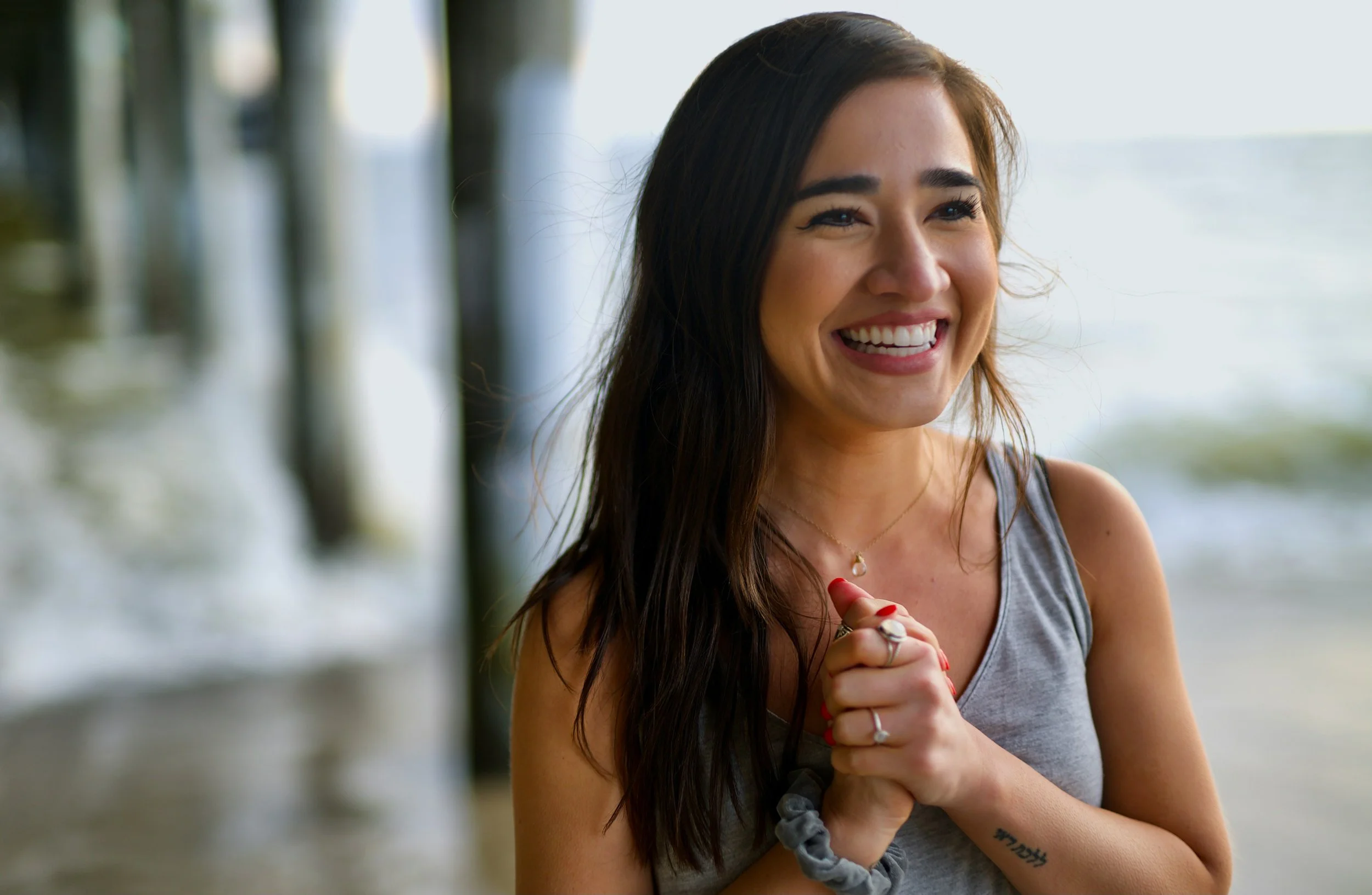 A woman with long dark hair smiling and holding her hands together near her chest on a beach with pier pillars and ocean in the background.