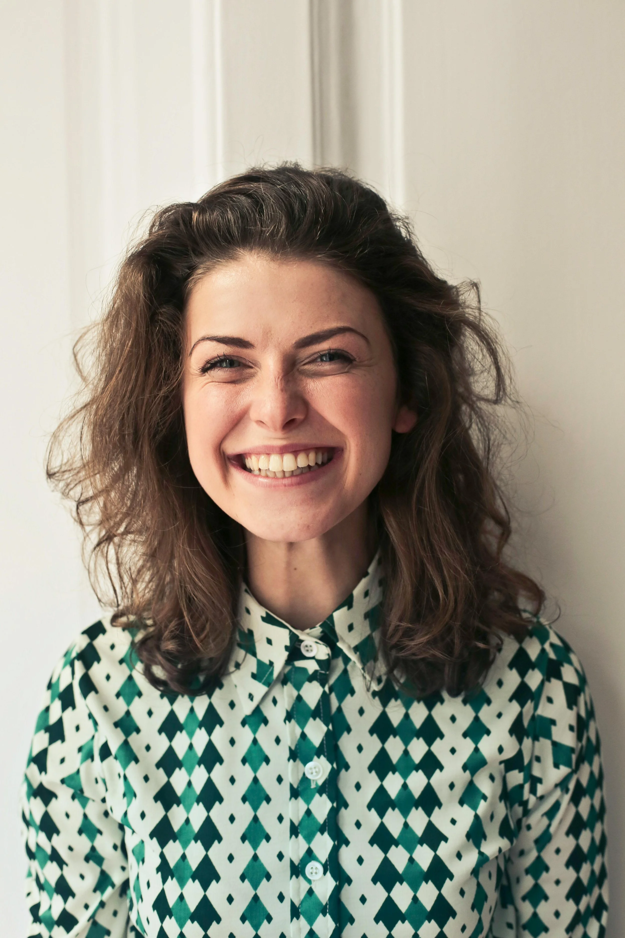 A woman with curly brown hair smiling widely, wearing a patterned button-up shirt, standing against a light-colored wall.