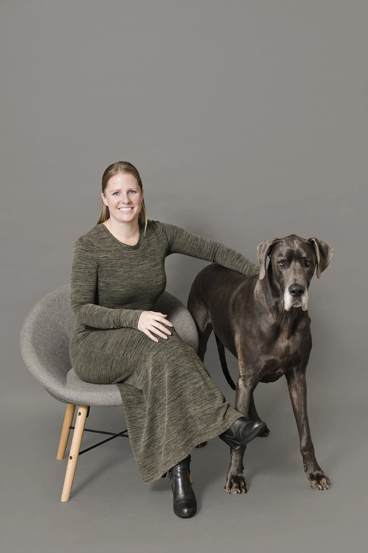 A woman sitting on a modern grey chair next to a large gray dog, both looking at the camera against a plain grey background.