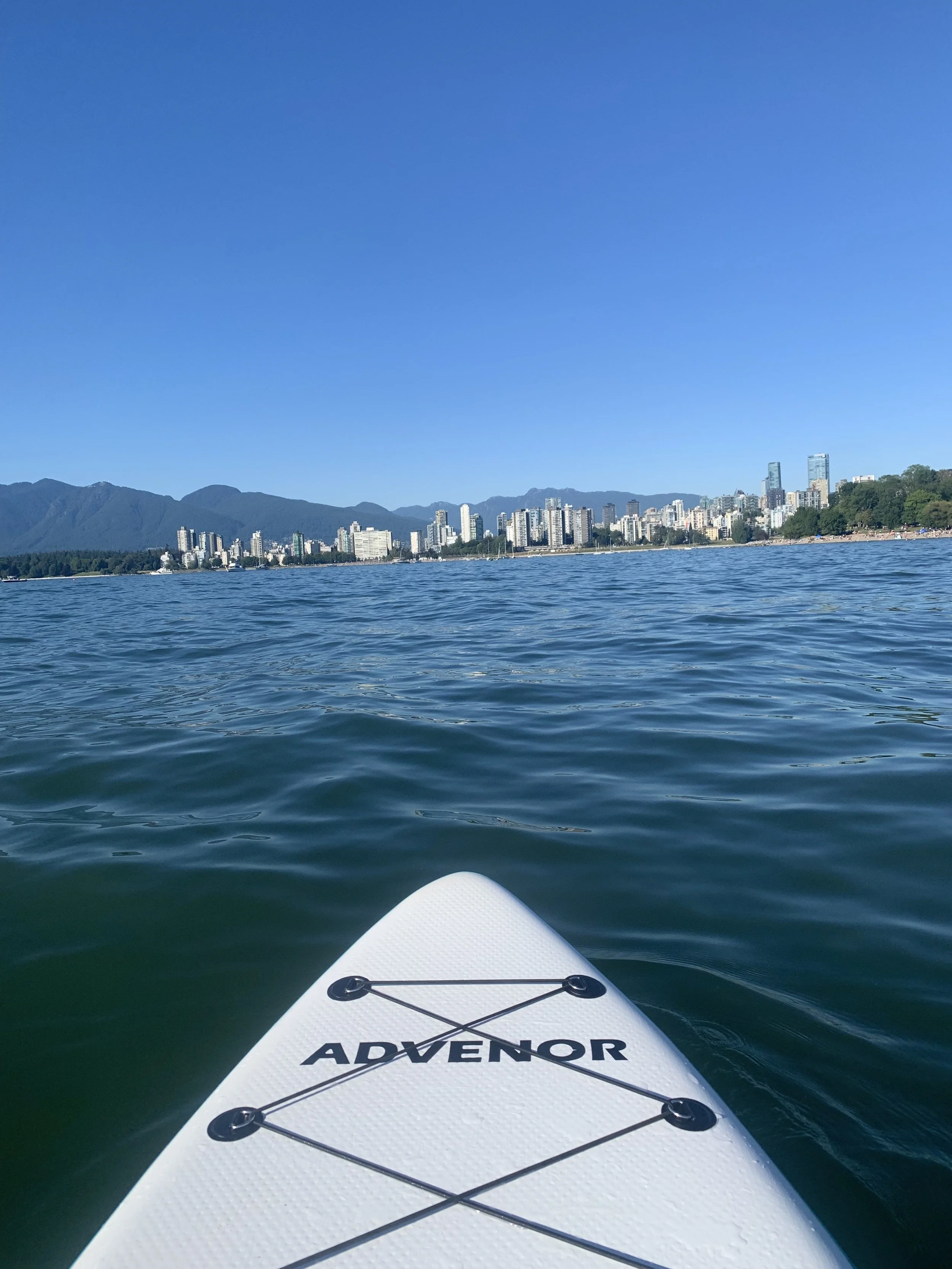 View from a paddleboard on the water, with a city skyline and mountains in the background.