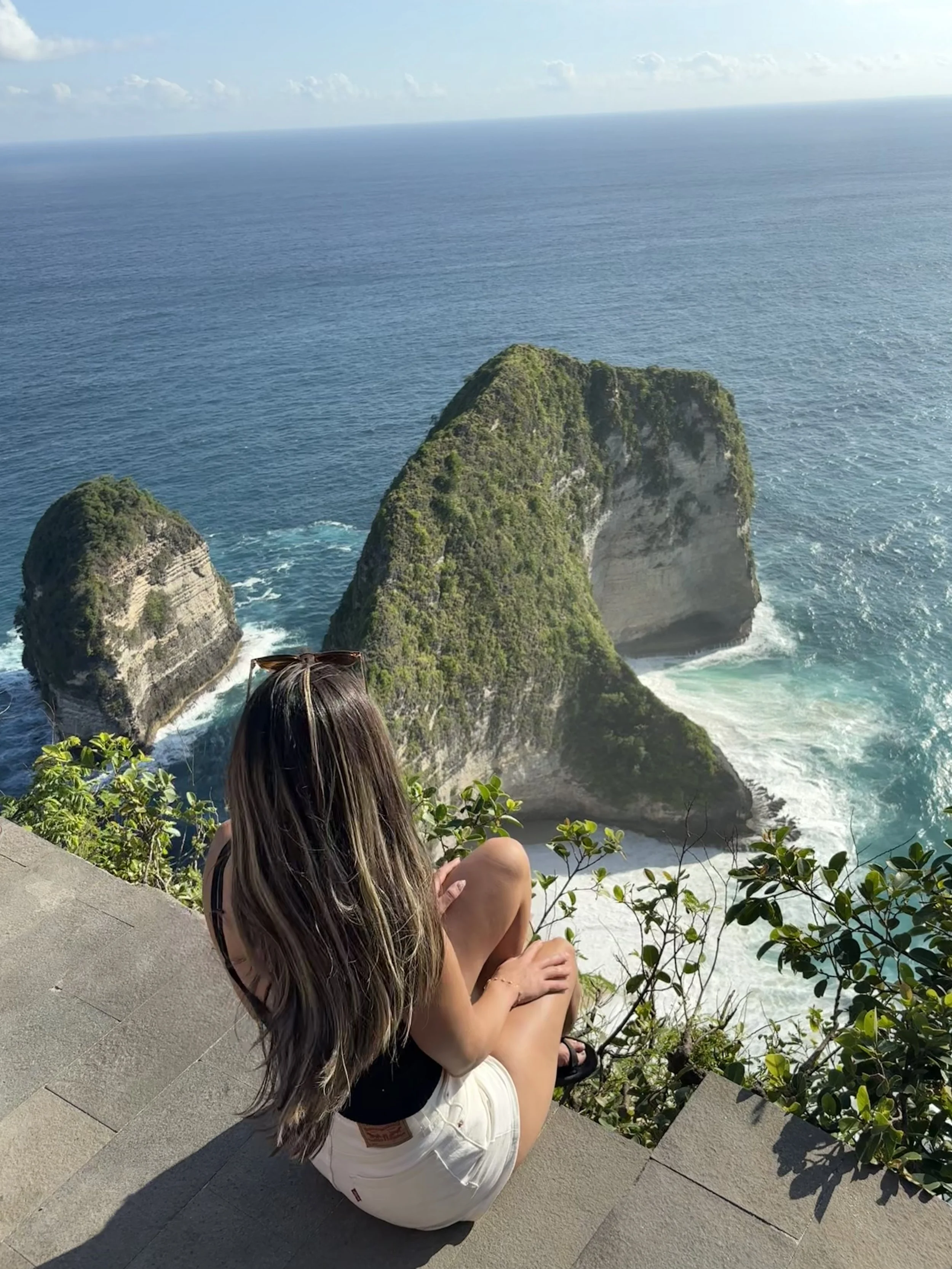 A woman sitting on a stone ledge overlooking large green-covered rock formations in the ocean, with blue sky and distant horizon.