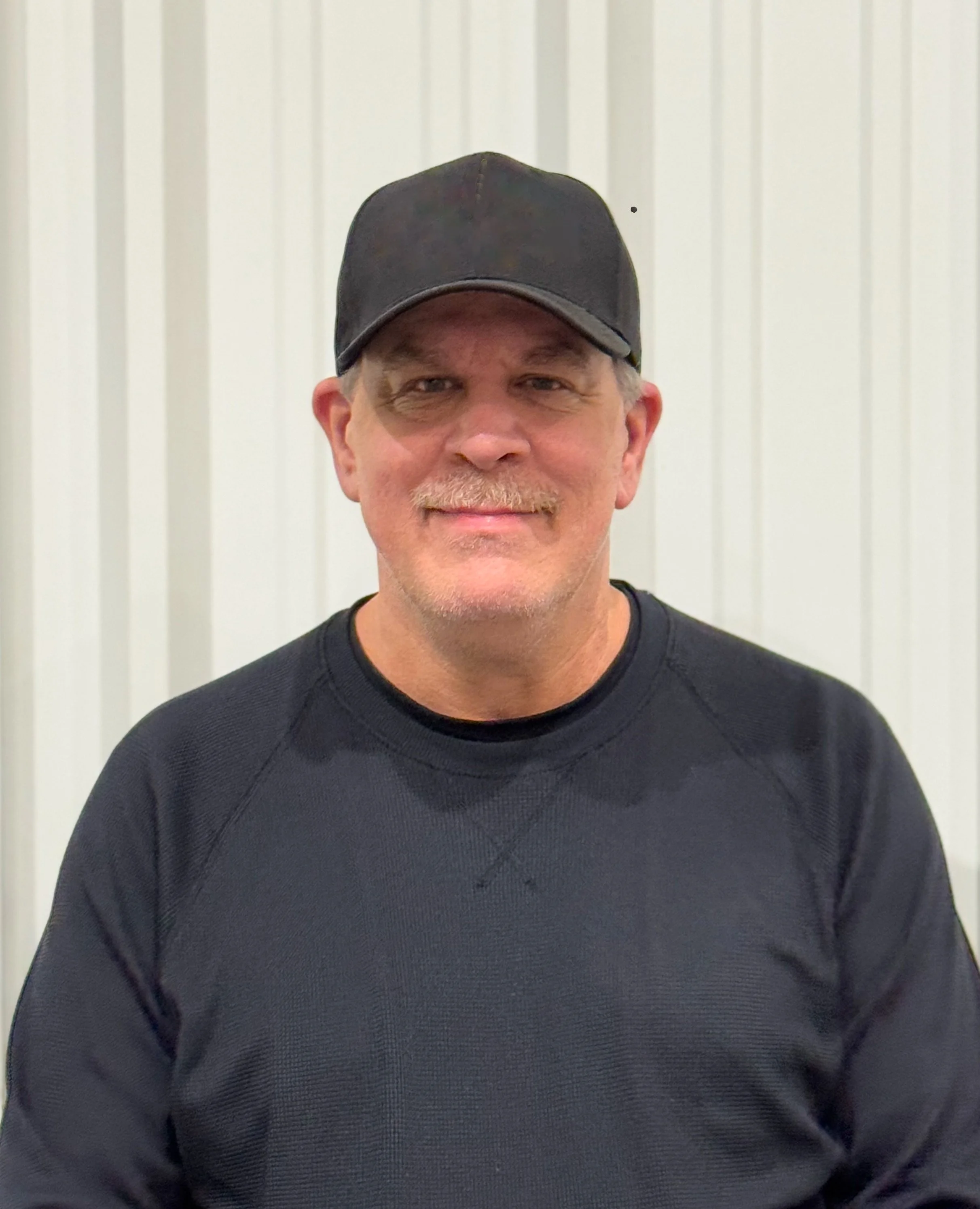 A middle-aged man with a mustache and gray hair, wearing a black baseball cap and black long-sleeve shirt, standing in front of a white paneled wall.