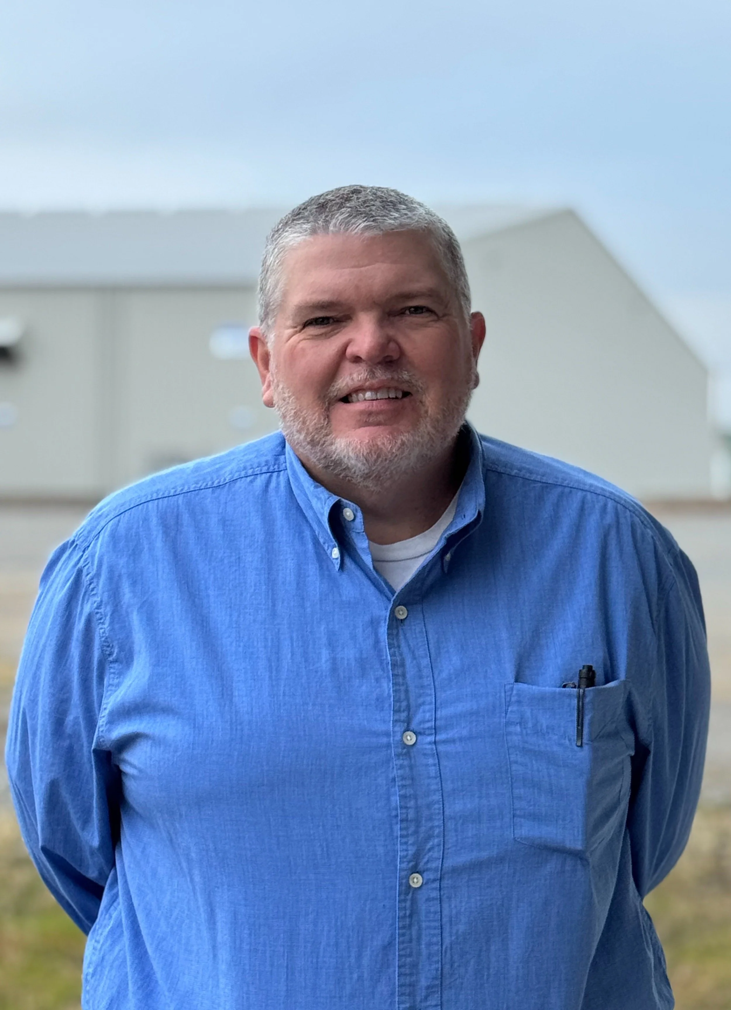 A middle-aged man with gray hair and beard, wearing a blue button-up shirt, standing outside with a building in the background.