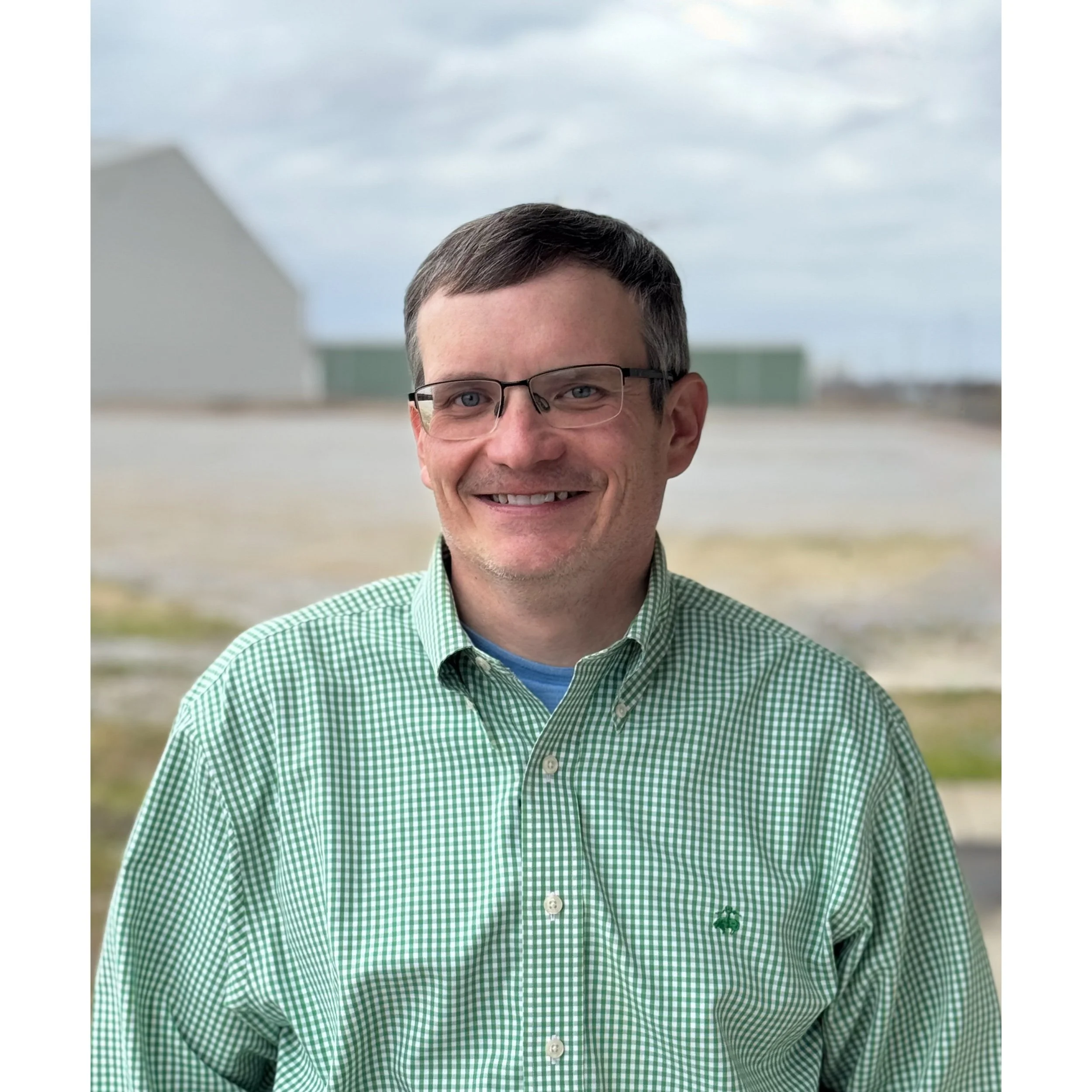 A man with glasses and short brown hair, smiling, wearing a green checkered shirt, standing outdoors with industrial buildings and a cloudy sky in the background.